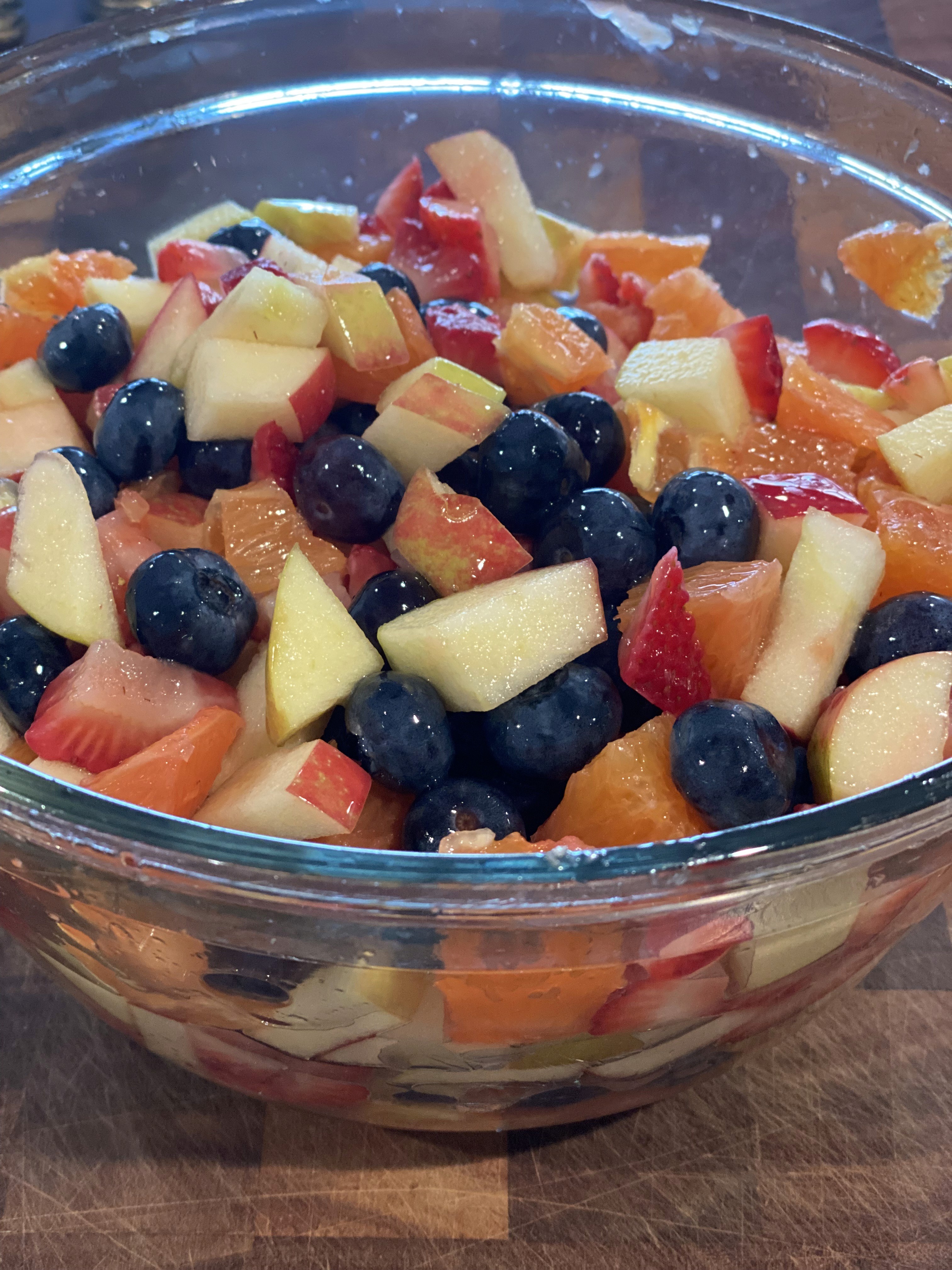 A clear glass bowl filled with a vibrant fruit salad, featuring a mix of diced apples, strawberries, blueberries, and orange segments, placed on a wooden surface.