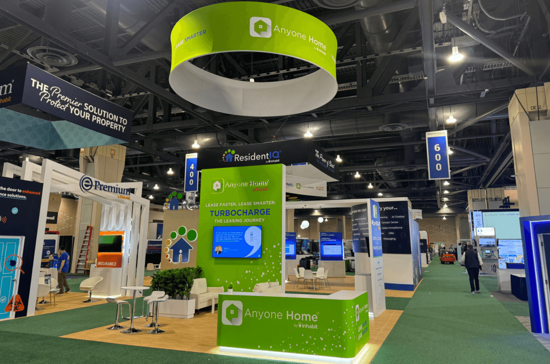 Anyone Home 20x20 professional service booth at NAA 2024 showing aisle view, green branded counter, tall software solution display, lounge seating, and neighboring service booths.