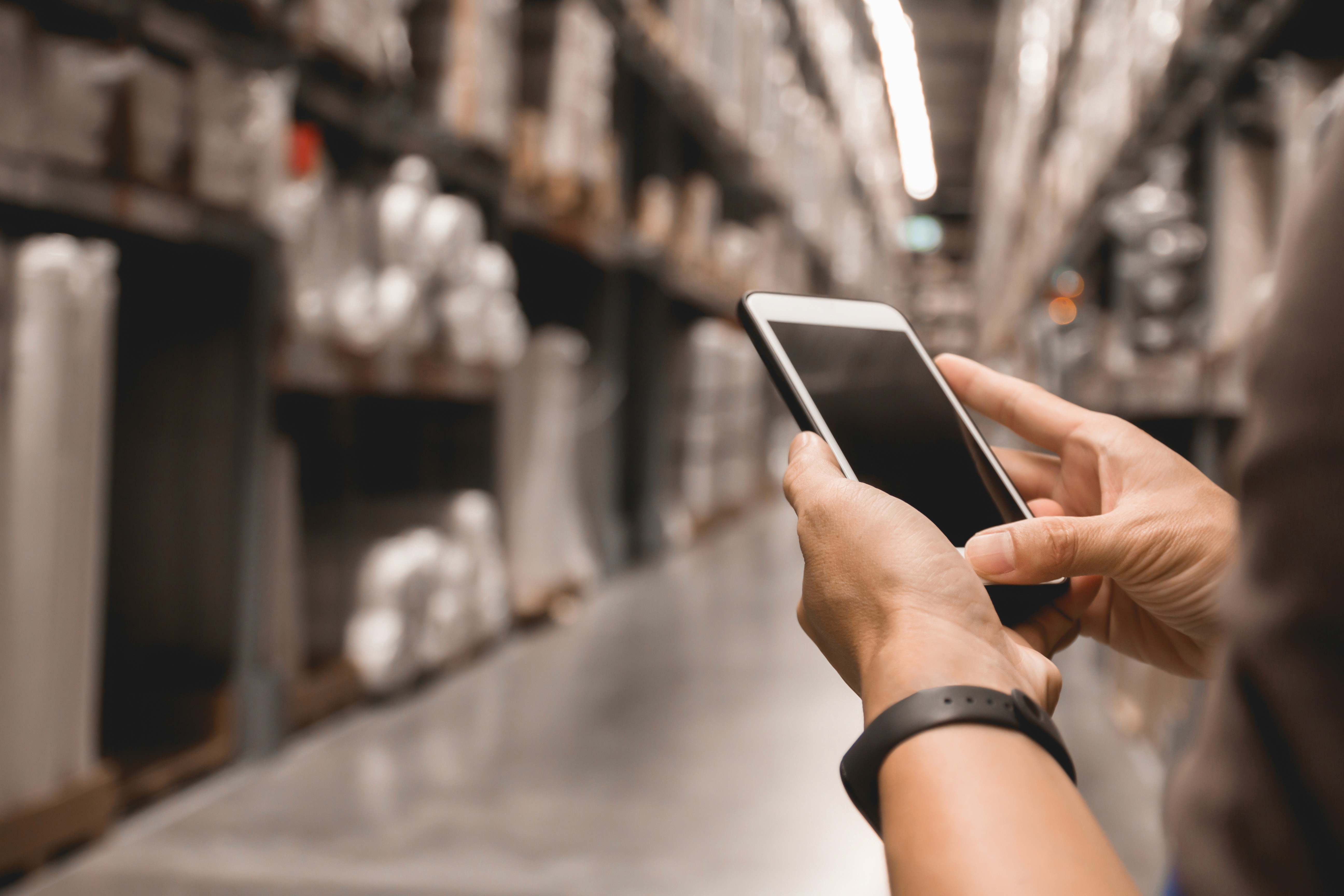 A person wearing a dark wristband is holding and using a smartphone in a large warehouse aisle, surrounded by shelves stocked with various goods, under bright industrial lighting.