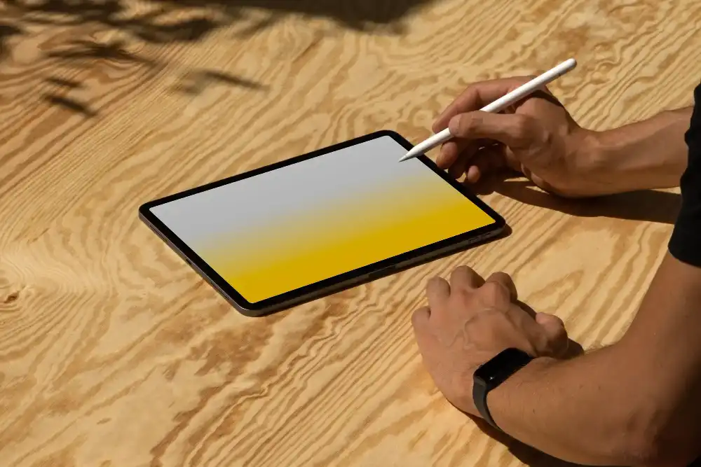 iPad mockup on a light wood surface, with a hand using a stylus and another resting nearby, wearing a smartwatch. The screen features a soft gradient background, emphasizing a modern, creative workspace.