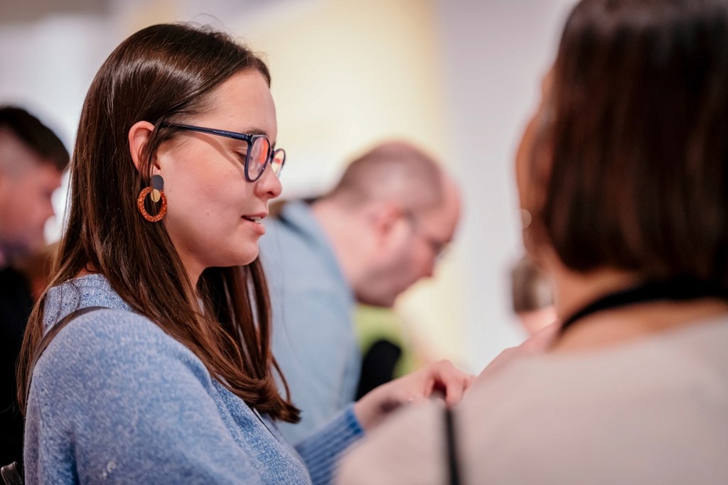 Several people standing and talking in an indoor event or gallery setting. In the foreground, one person with long brown hair is wearing a blue jumper and circular orange earrings, turned slightly toward another person with short dark hair and a light‑colored top. Other attendees are visible in the background, slightly out of focus, creating a sense of a busy, social atmosphere.