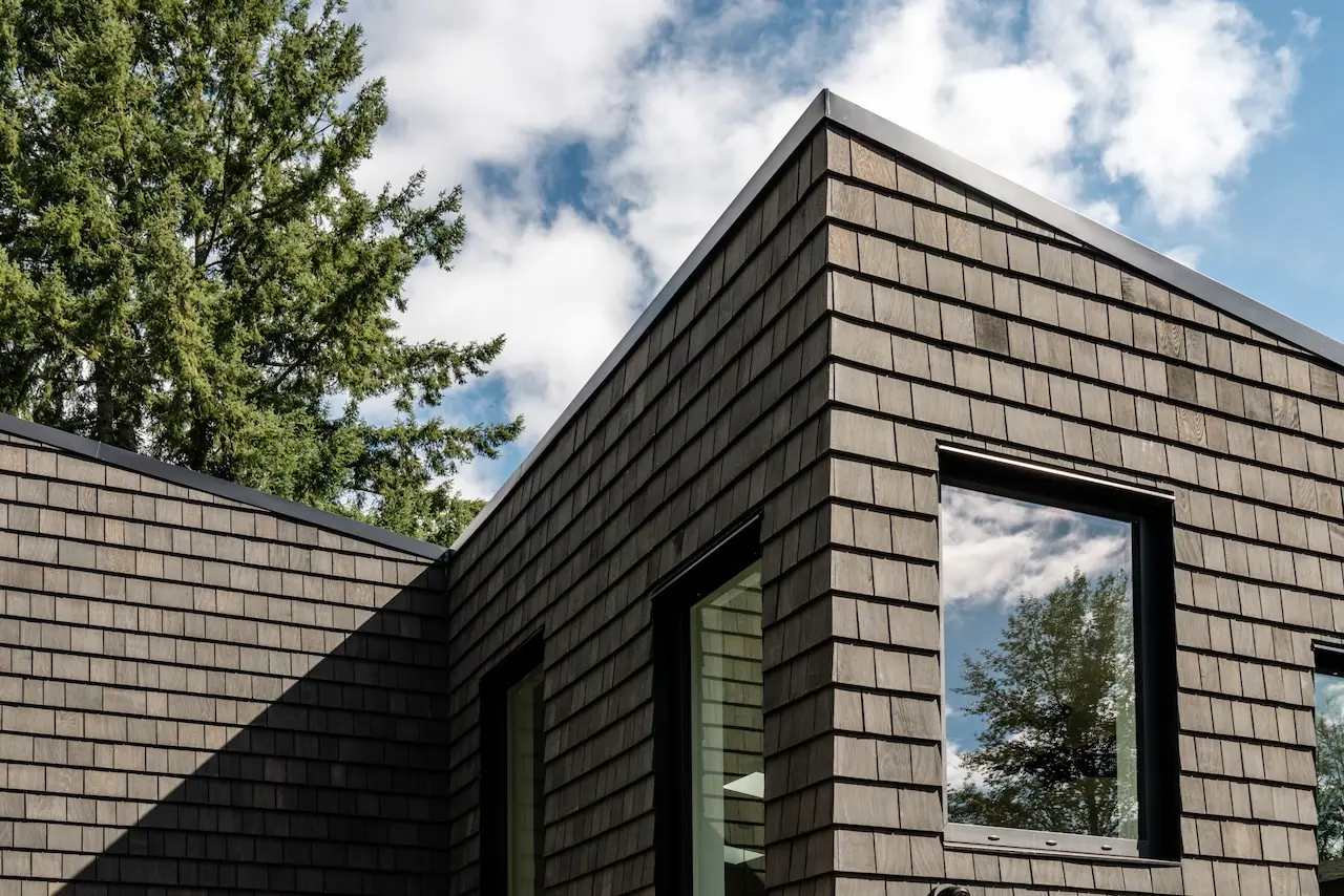 Modern two-story home with large glass windows, warmly lit interiors, and a balcony. Surrounded by trees and a manicured lawn at dusk. Cozy ambiance.
