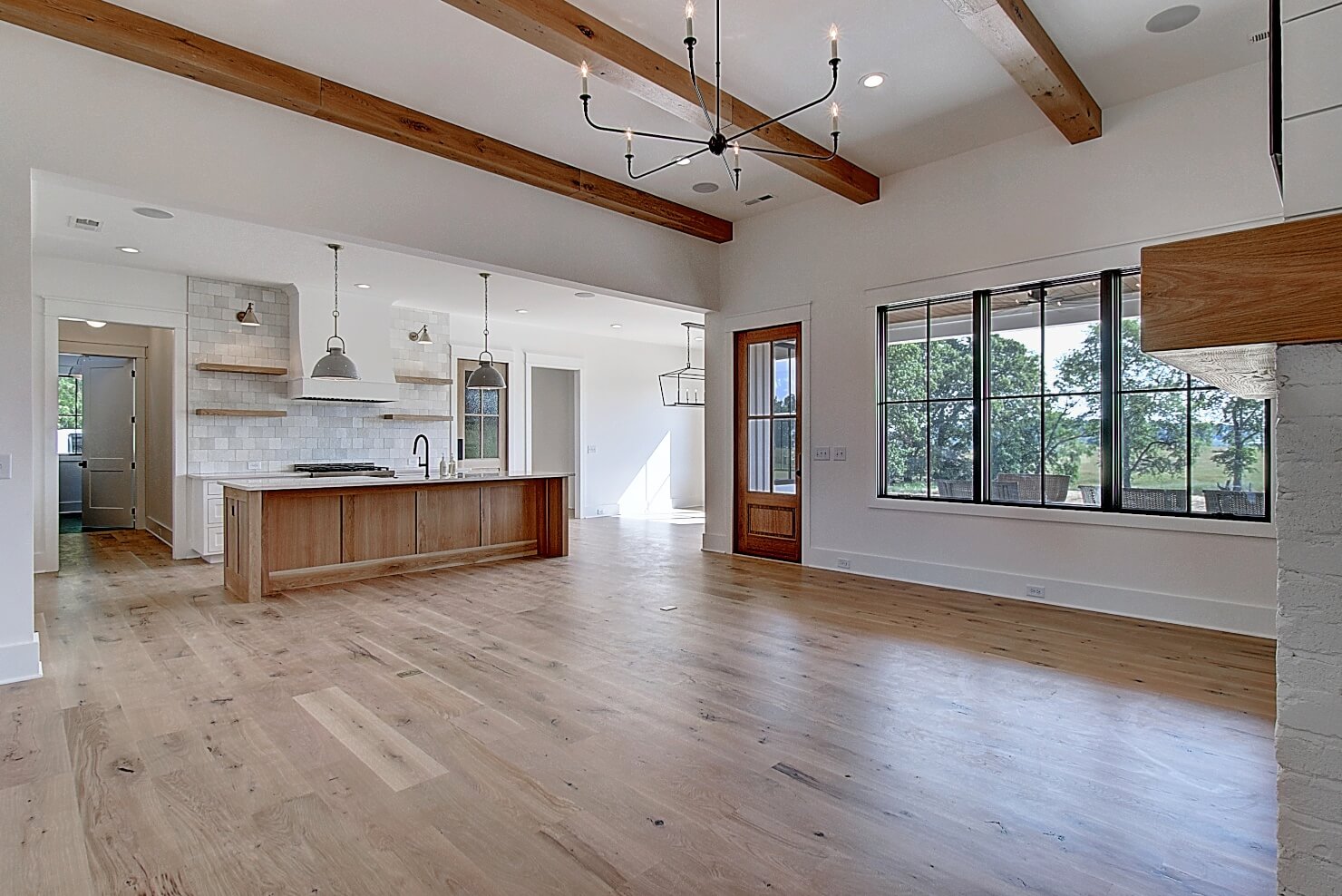 Modern open-concept living space with wooden beams, large windows, and a kitchen area featuring wood cabinetry.