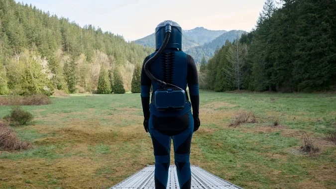 A person in a blue and black futuristic suit stands on a metal platform, overlooking a lush, tree-lined landscape with mountains in the background, under a clear sky.