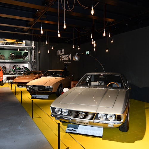 A showroom with vintage cars displayed under hanging lights, featuring a “ITALO DESIGN” sign in the background.