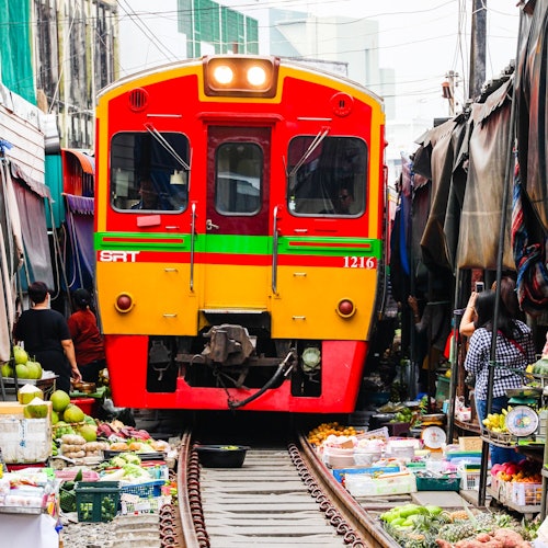 붉고 노란색의 기차가 양쪽에 여러 농산물과 상품을 가득 싣고 있는 좁은 시장을 통과합니다.