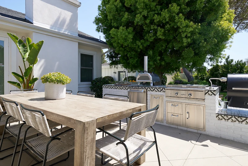 Sunny outdoor dining area with a rustic table set, white built-in BBQ, lush tree backdrop, and potted plants. Photo by Todd Huge.