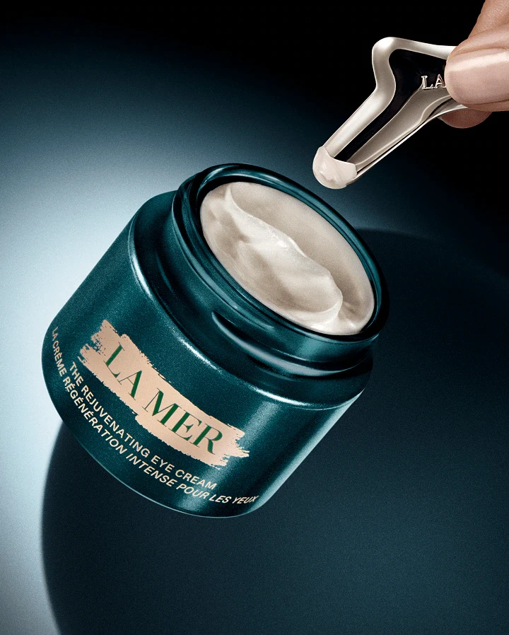 A close-up of La Mer rejuvenating eye cream jar, with a small applicator tool about to scoop out the creamy product.