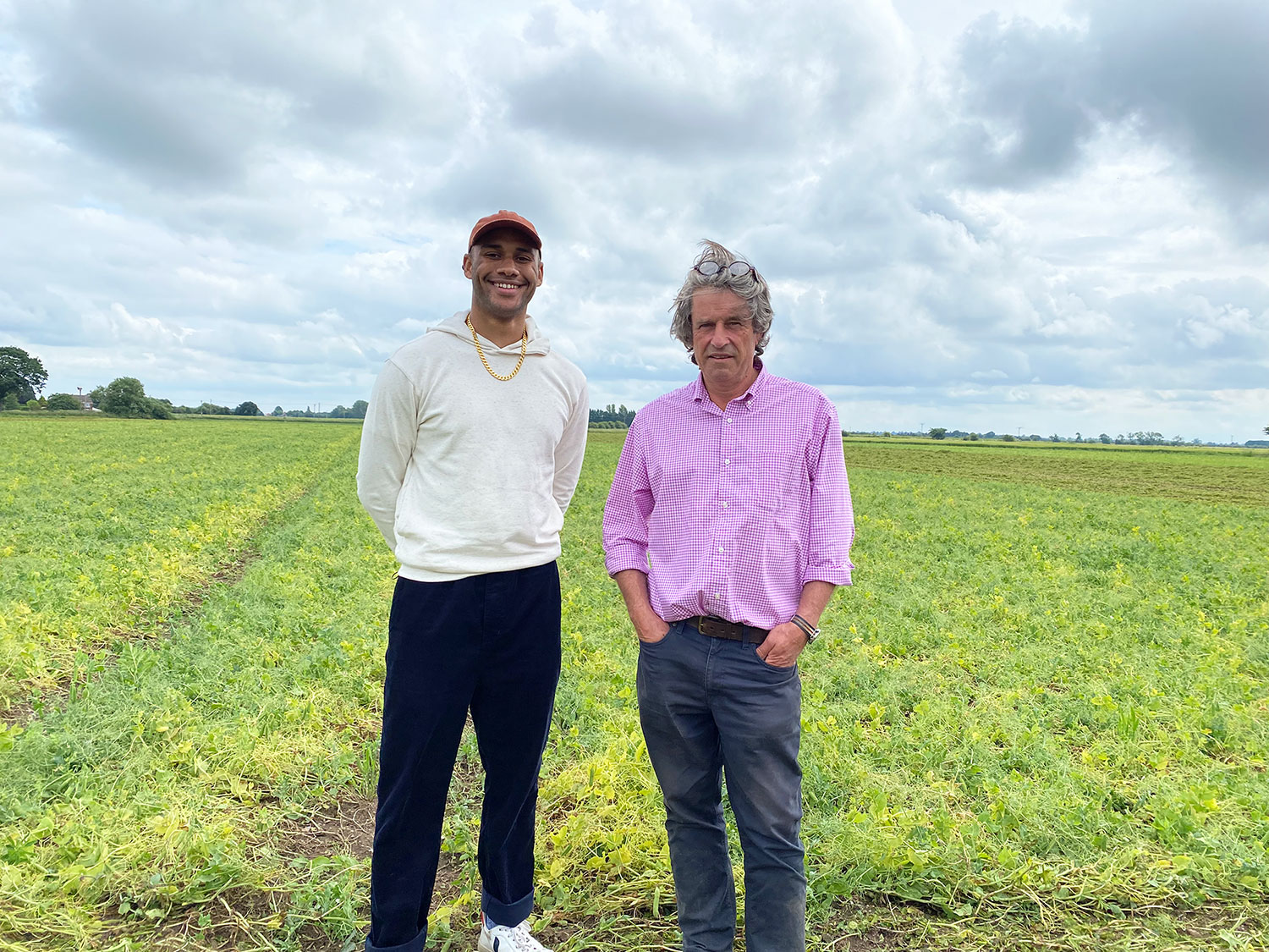 Joe and Stephen standing in a field of green peas. Joe is wearing a cap and a pale sweatshirt. Stephen is wearing a pink shirt with grey trousers