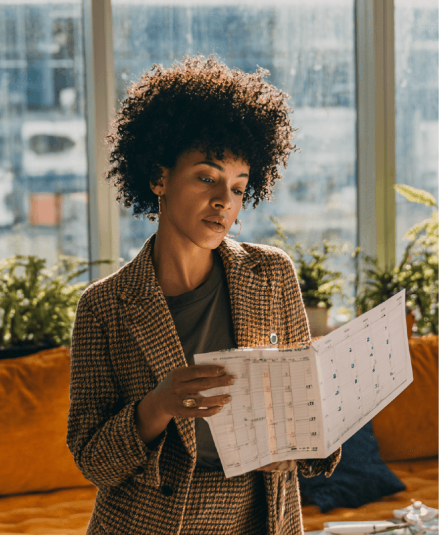 Profissional em escritório analisa um calendário impresso de prazos de convênios, com plantas e vista urbana ao fundo; organização financeira de clínicas na Tivita.