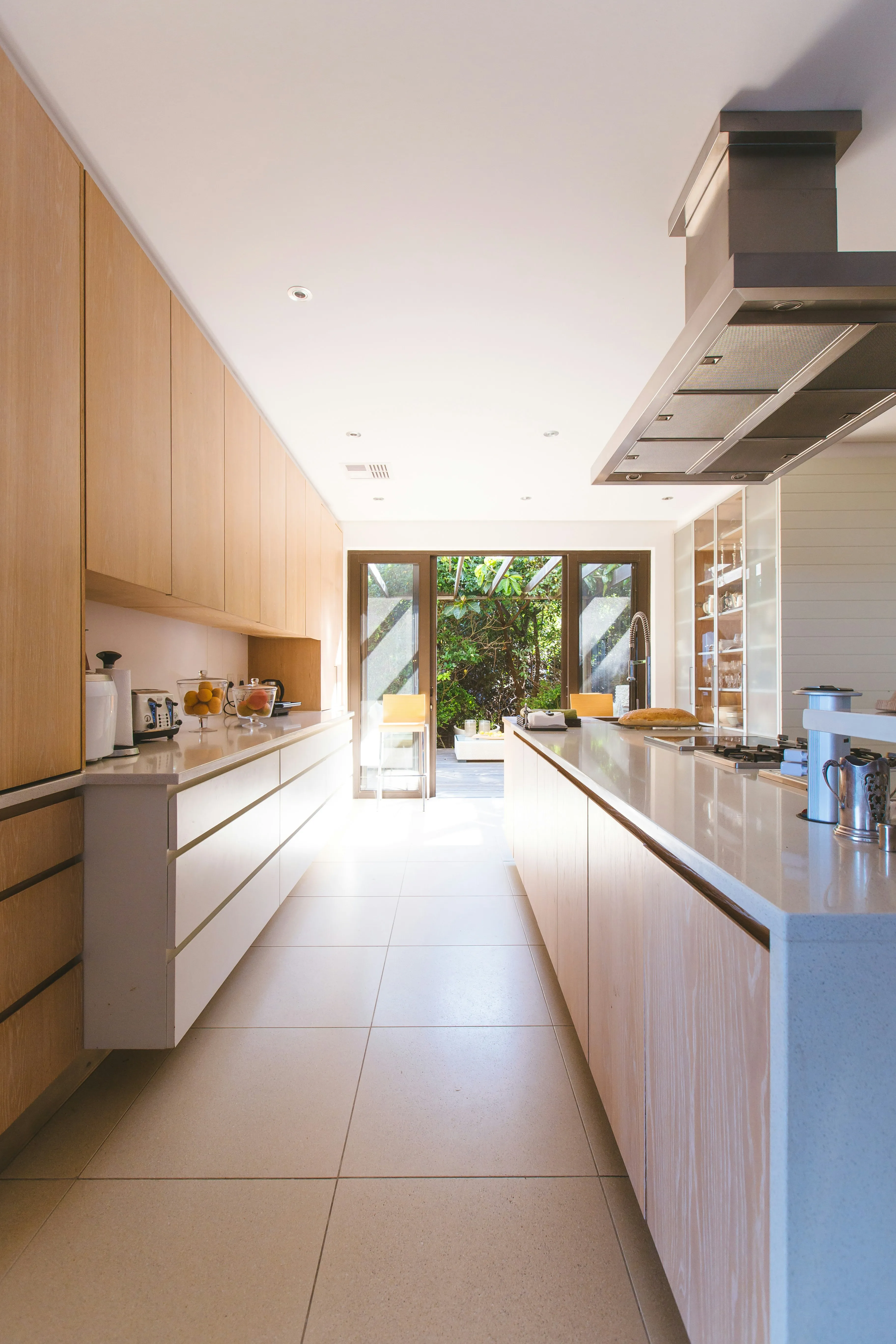 Modern kitchen with light wood cabinetry, sleek countertops, and a view of greenery through large glass doors.