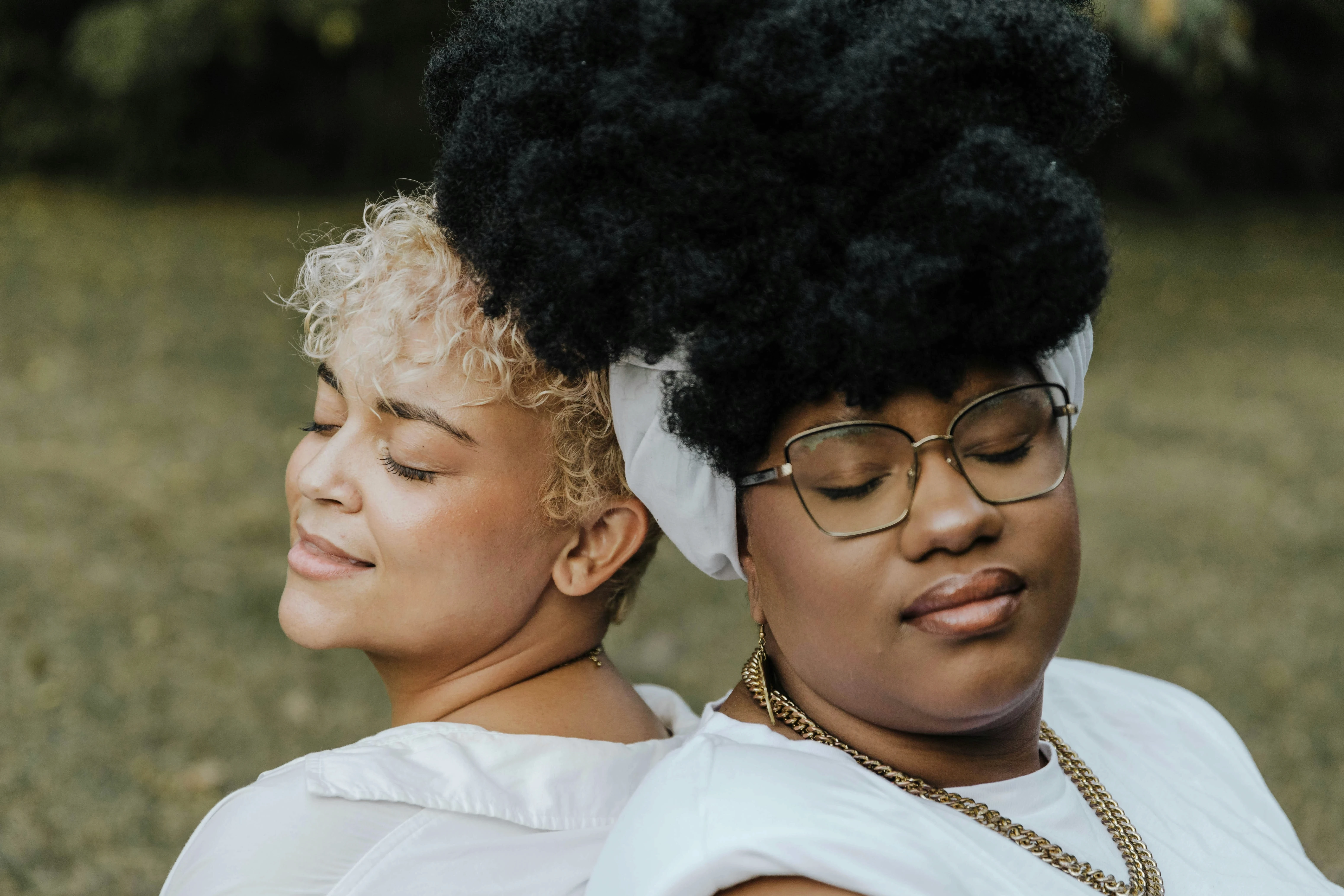 Two adult women standing back-to-back outdoors with their eyes closed, conveying calm, connection, and self-assurance.