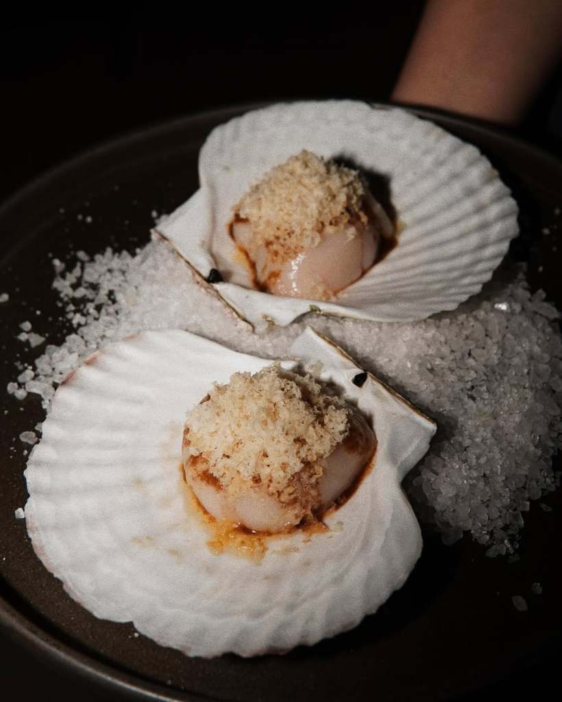 A close-up of two scallops presented in their shells on a bed of salt, topped with a crispy garnish.