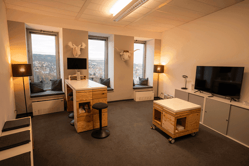 Modern eingerichteter Raum mit drei Fenstern, zwei Stehlampen, einem hohen Tisch mit Hockern, einem rollbaren Holztisch und einem Fernseher auf einem Sideboard