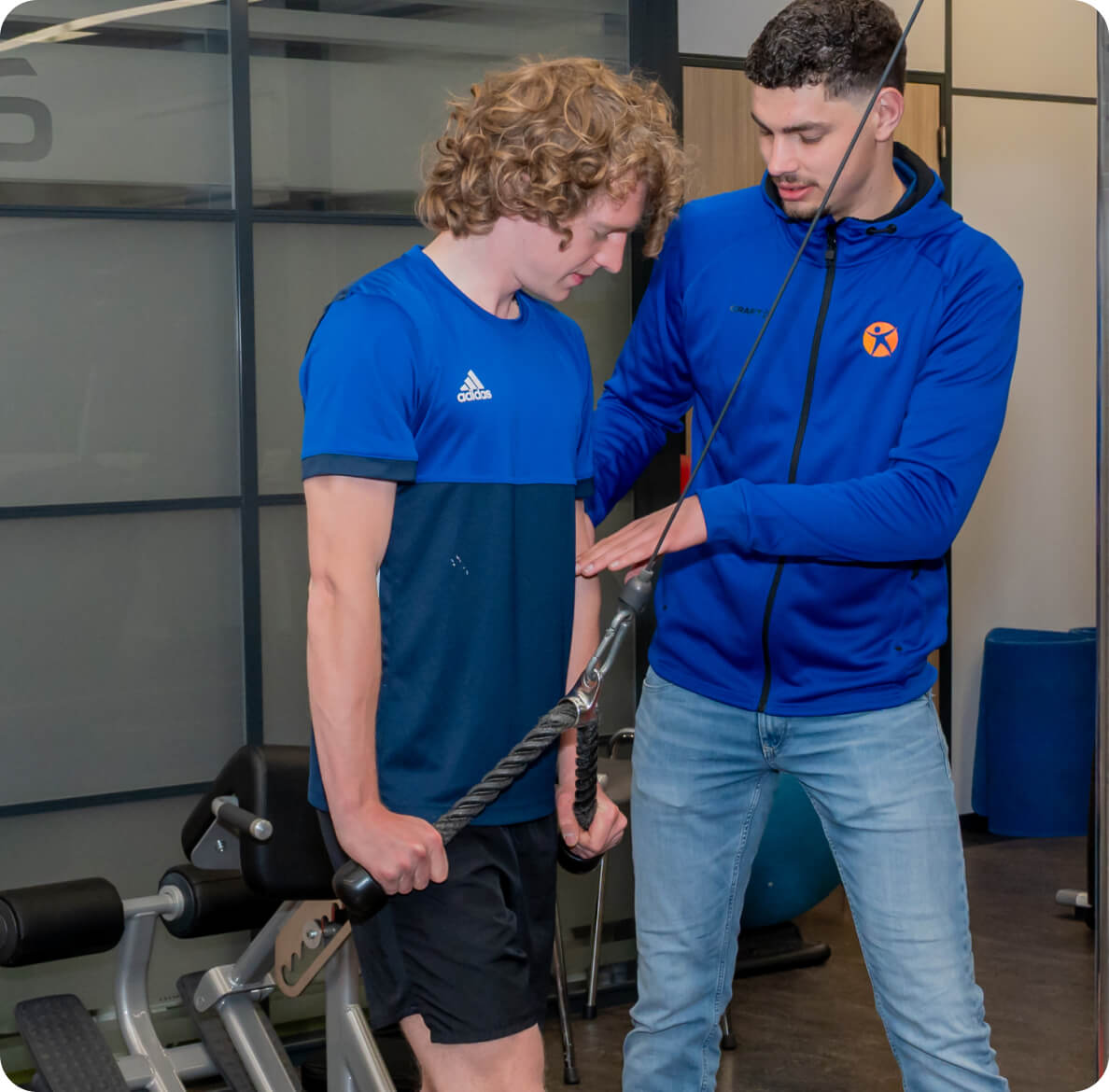 A personal trainer wearing a blue hoodie is instructing a man in a blue athletic shirt on proper cable machine exercise form at the gym.