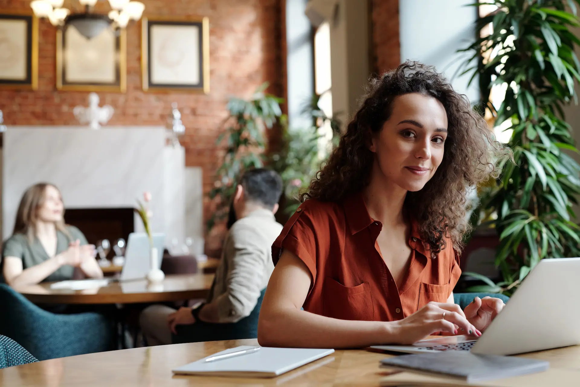 A woman with curly hair wearing a rust-colored blouse is sitting at a wooden table with a laptop and notebook, in a cozy, plant-decorated cafe with exposed brick walls and framed artworks.