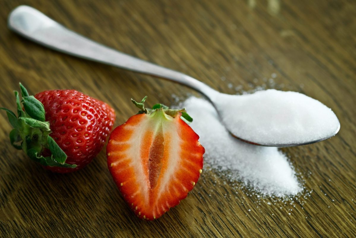 Some strawberries sit on the counter next to a spoonful of sugar.
