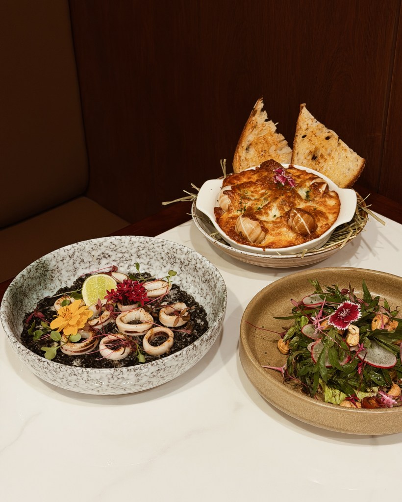 A beautifully arranged meal featuring three dishes: a bowl of black squid ink pasta topped with sliced squid and garnished with edible flowers, a baked dish with a golden crust and herbs, accompanied by slices of toasted bread, and a salad with greens, nuts, and colorful floral accents.