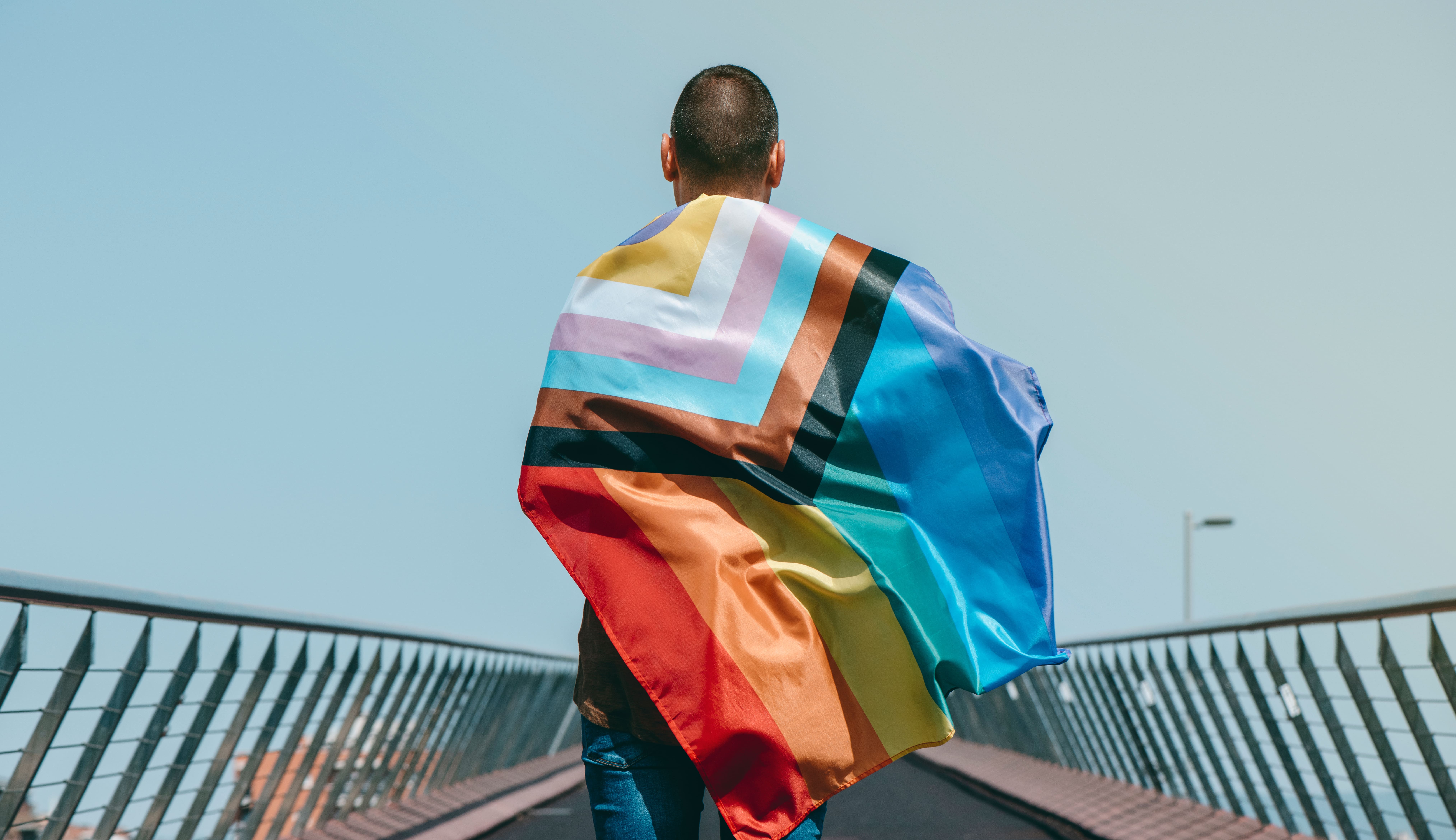 A imagem mostra uma pessoa de costas, vestindo uma bandeira progressista do orgulho LGBTQIA+. A bandeira apresenta um design com várias cores, incluindo as tradicionais do arco-íris, além de tons que representam pessoas trans, negras e marginalizadas dentro da comunidade. A pessoa está caminhando em uma ponte sob um céu claro, e a perspectiva sugere um movimento em direção ao futuro, destacando a mensagem de inclusão e progresso.