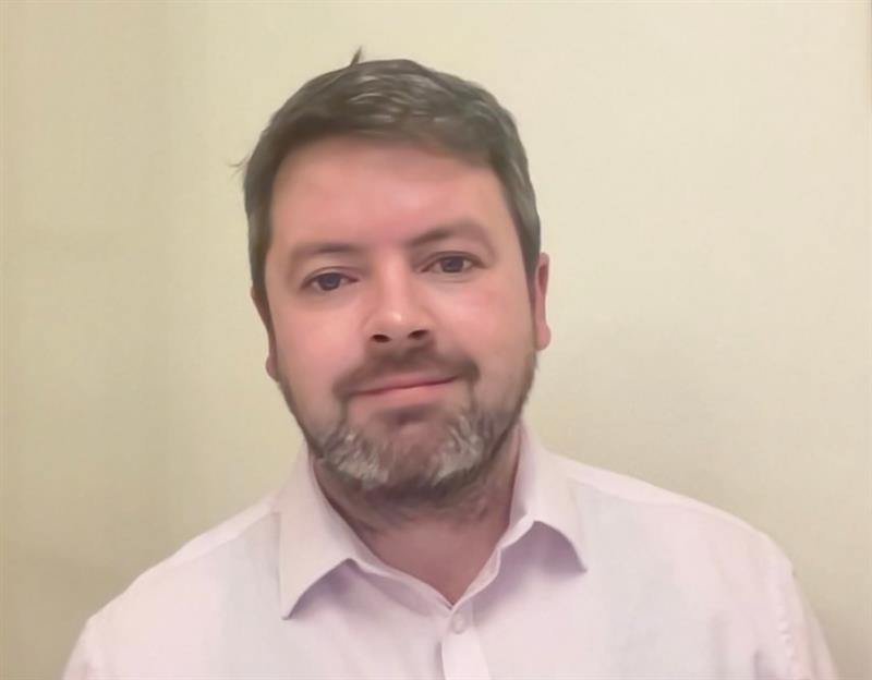 A person shown from the chest up, wearing a light-colored button-down shirt with an open collar, standing against a plain, light-colored wall. The image is evenly lit with a neutral indoor background and no visible objects or text.