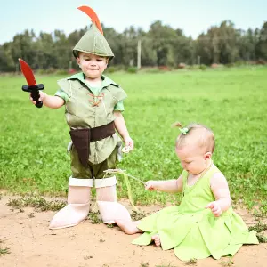 שני ילדים קטנים מחופשים בטבע לרגל חג פורים: פעוט בתחפושת פיטר פן אוחז בחרב צעצוע ותינוקת בתחפושת פיה ירוקה יושבת על הקרקע. צילום ילדים אמנותי באור יום מאת הצלמת שירי עציץ Shiriz Photography