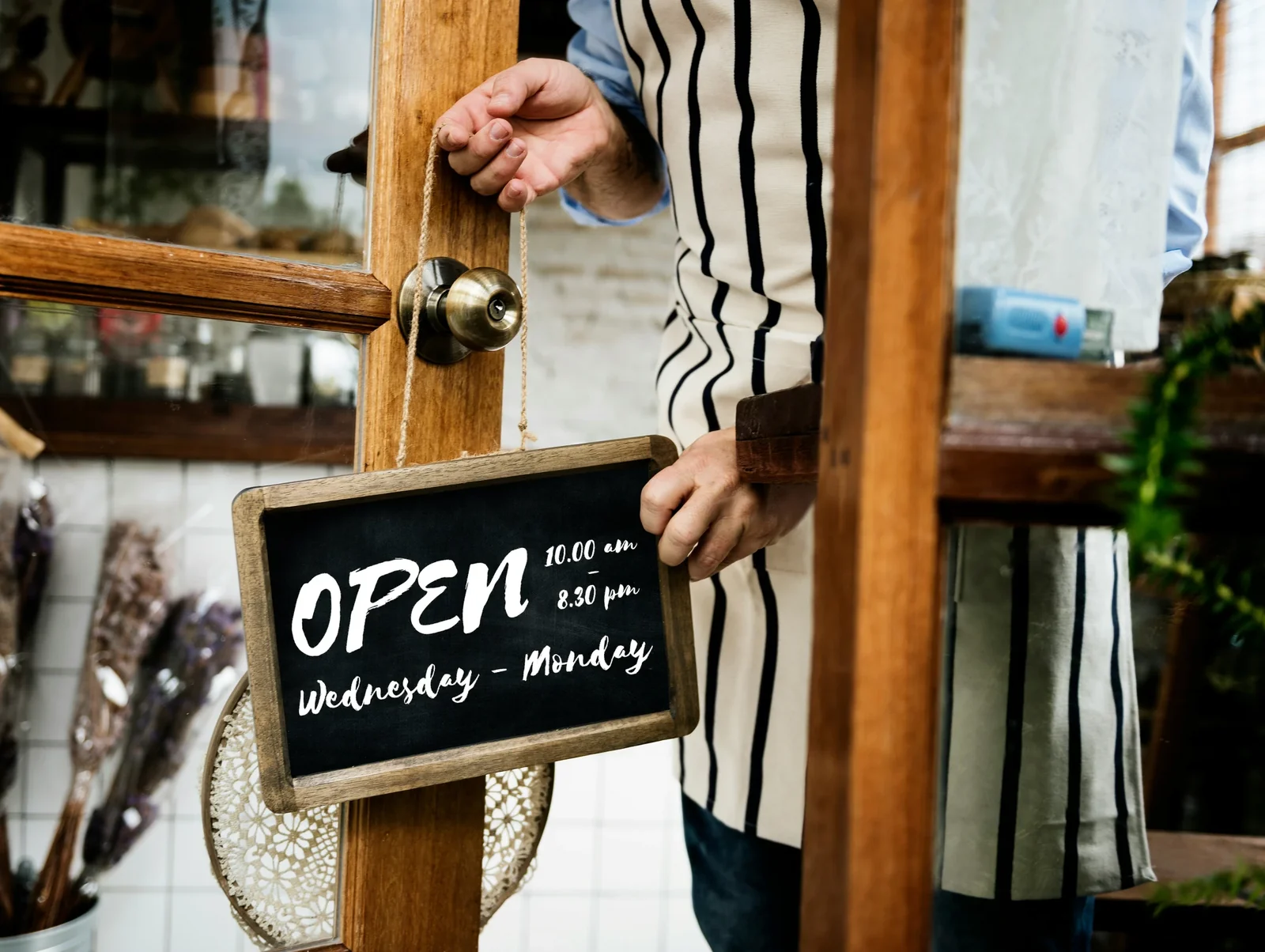 Person holding "OPEN 10:00 am 8:30 pm Wednesday - Monday" sign on wooden door with brass knob.