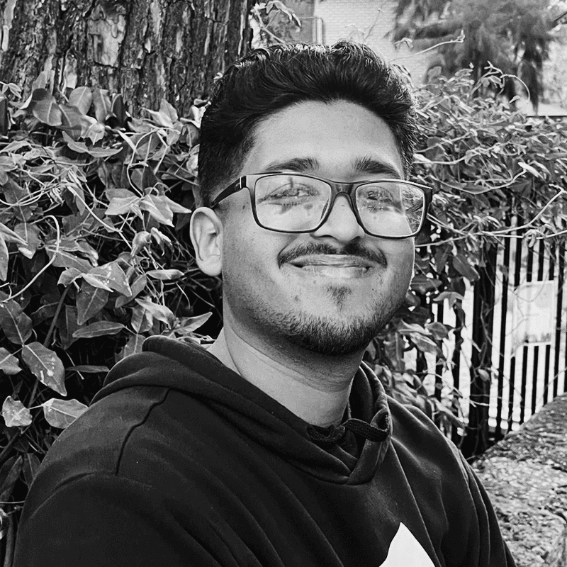 A woman sitting at a desk in a modern office, working on a laptop with documents and plants around her. Sunlight filters through large windows in the background.