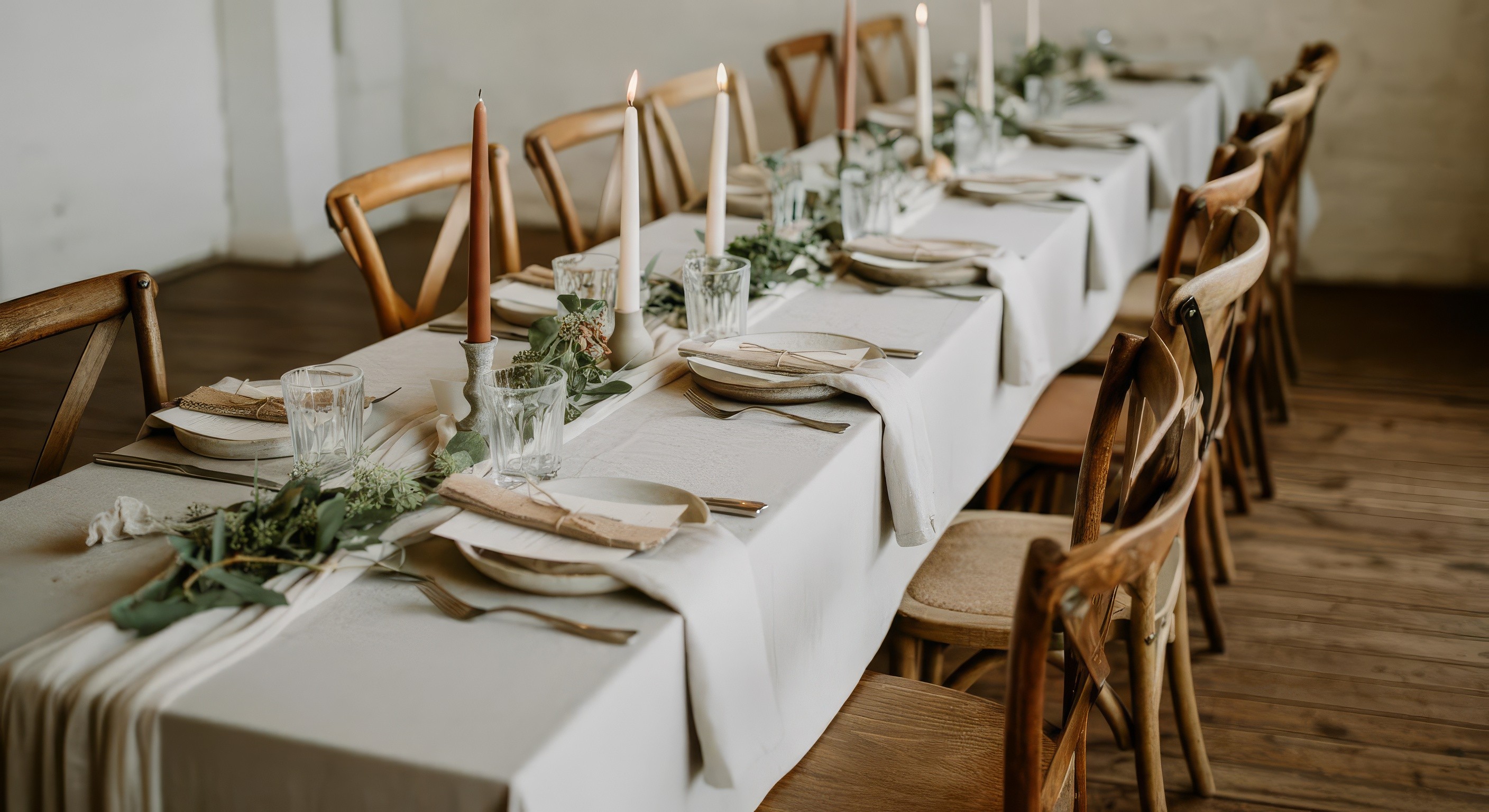 A beautifully set dining table with white tablecloth, cutlery, and fresh greenery in a cozy, well-lit space.