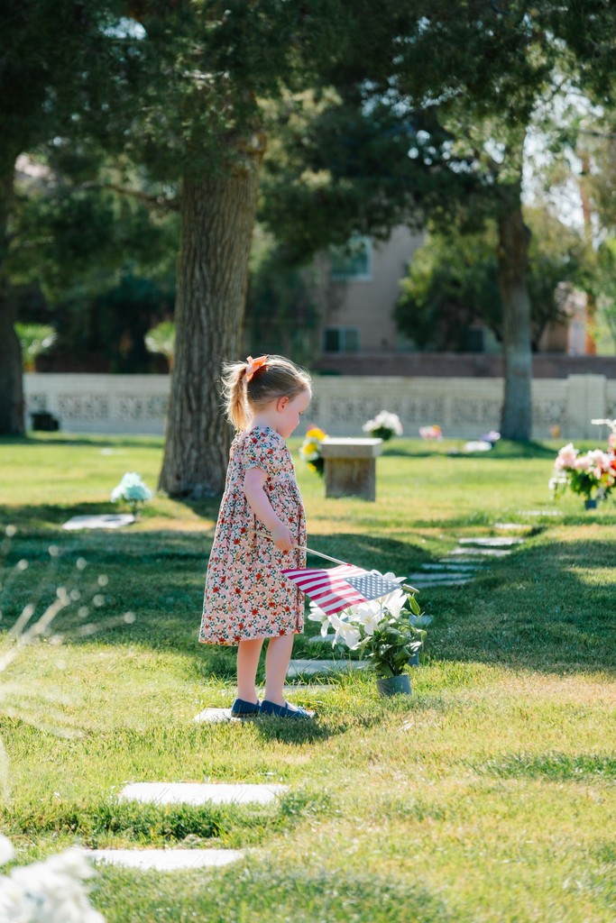 A young girl in a floral dress with a red bow stands solemnly in a cemetery, holding an American flag near a grave adorned with white flowers, paying tribute on a sunny day.