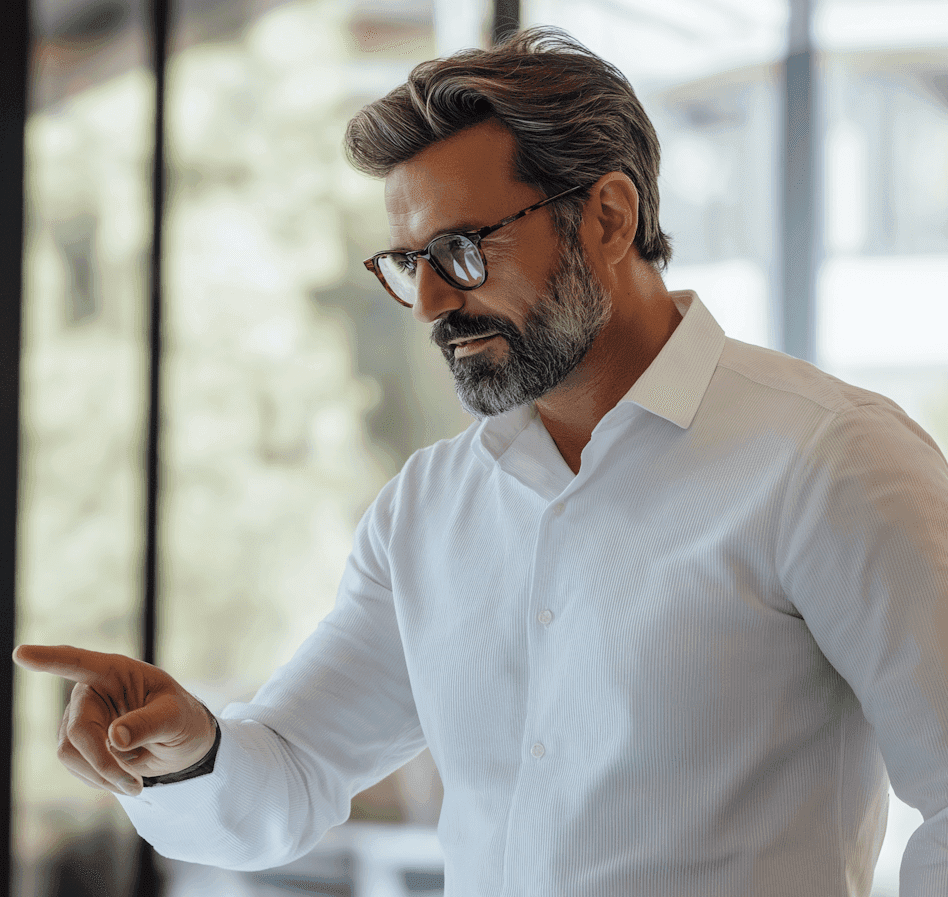 A bearded man wearing glasses points at an object, indicating interest or emphasis in a discussion.