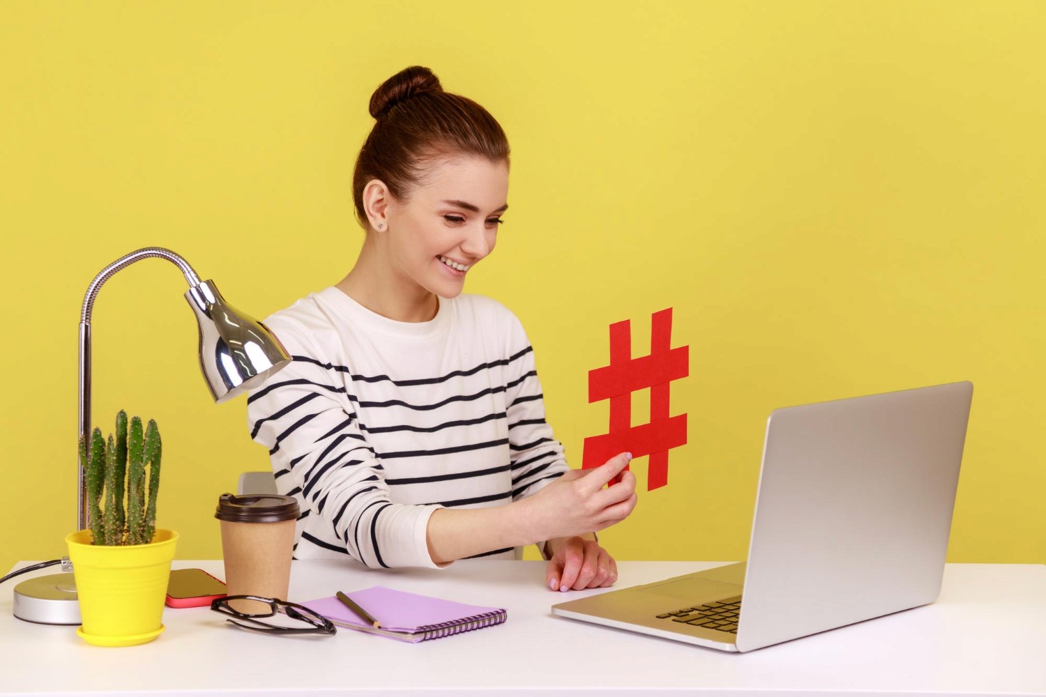 A woman holds a red hashtag symbol in front of her laptop, symbolizing social media engagement and digital communication.