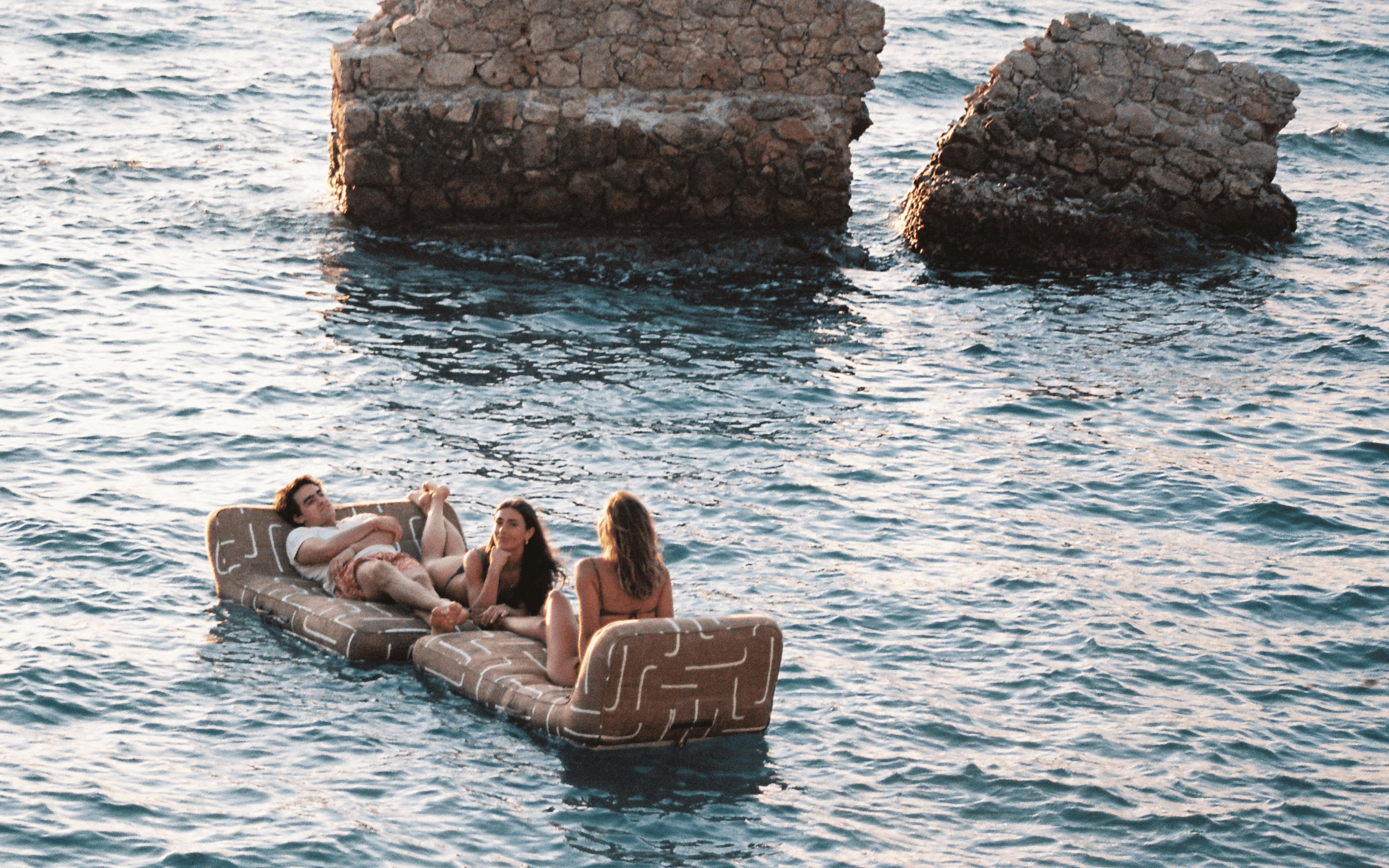 Three people lounging on a gold-and-brown textured luxury pool floats in the sea near rocky ruins