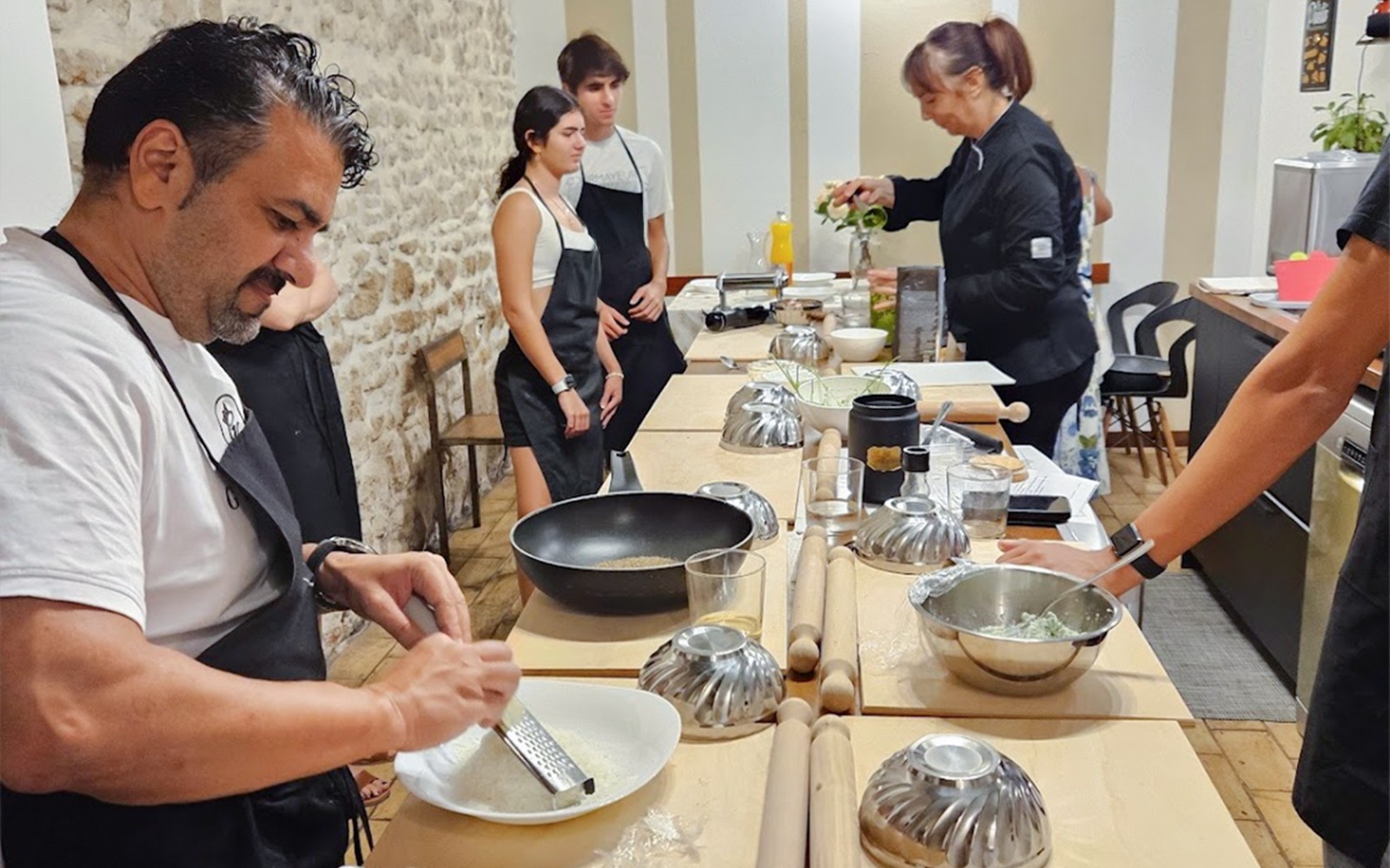 रोम में एक कुकिंग क्लास के प्रतिभागी एक रसोई काउंटर पर व्यंजन तैयार कर रहे हैं।