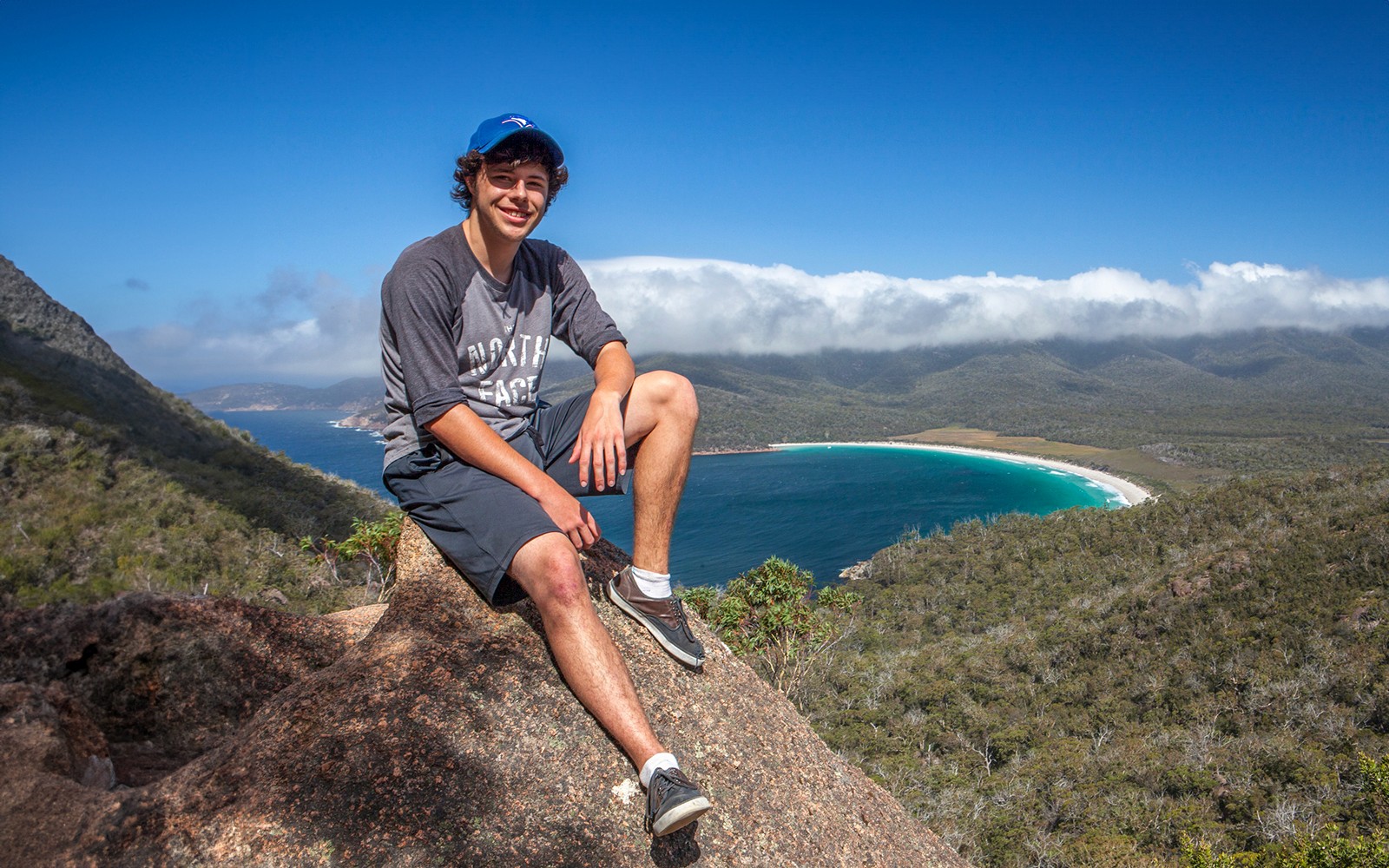Person sitting on rock overlooking Wineglass Bay, Tasmania, with ocean and forested hills in the background.