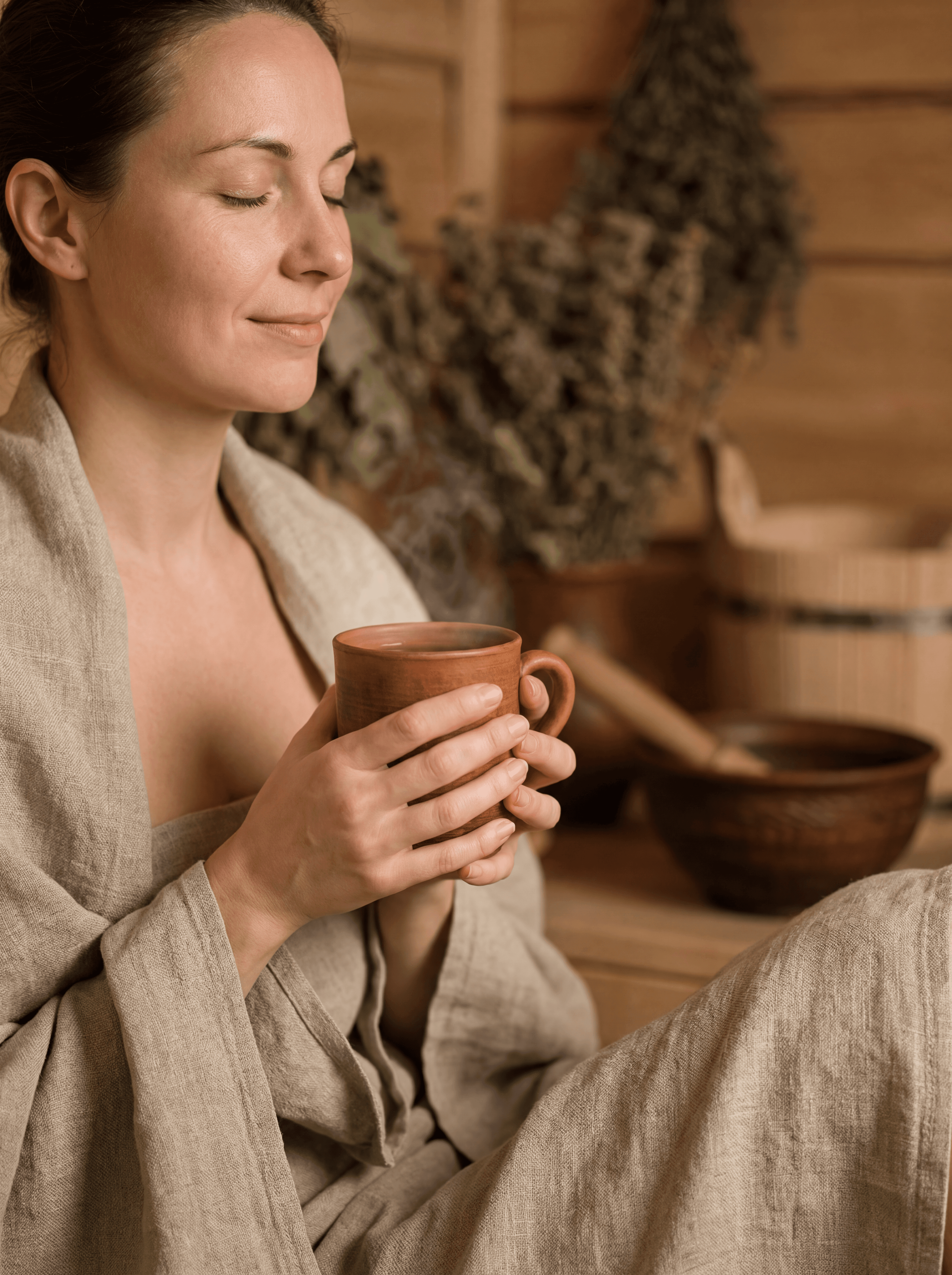A close-up, intimate indoor scene focused on women’s holistic wellness: a woman seated peacefully after treatment, wrapped in linen, holding a warm herbal infusion or resting hands on her abdomen. No overt medical cues. Soft lighting, blurred wooden interior background, gentle expression conveying emotional balance and vitality. Authentic Ayurvedic environment, minimal composition, nurturing and respectful mood. Elegant, natural wellness aesthetic.