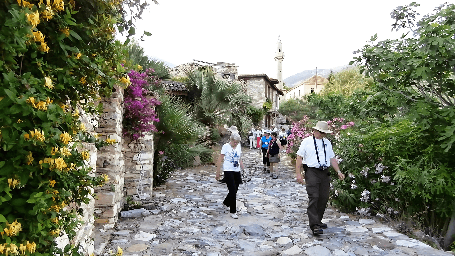 Travelers walking along a historic stone street lined with lush gardens and Ottoman houses in a traditional Turkish village