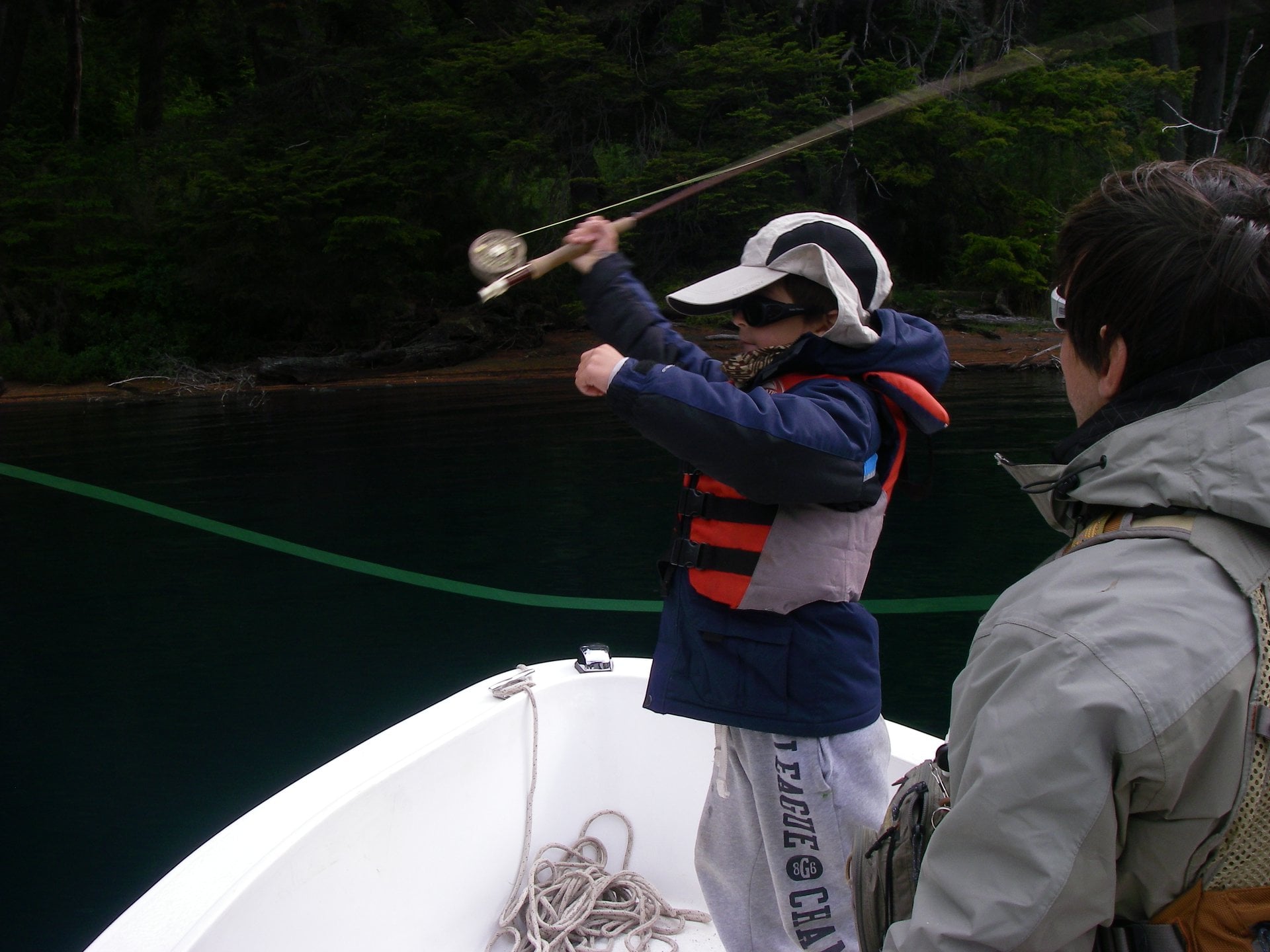 Niño blandiendo su caña hacia atrás, mientras la linea dibuja una figura en el aire. El niño esta sobre un bote en el río