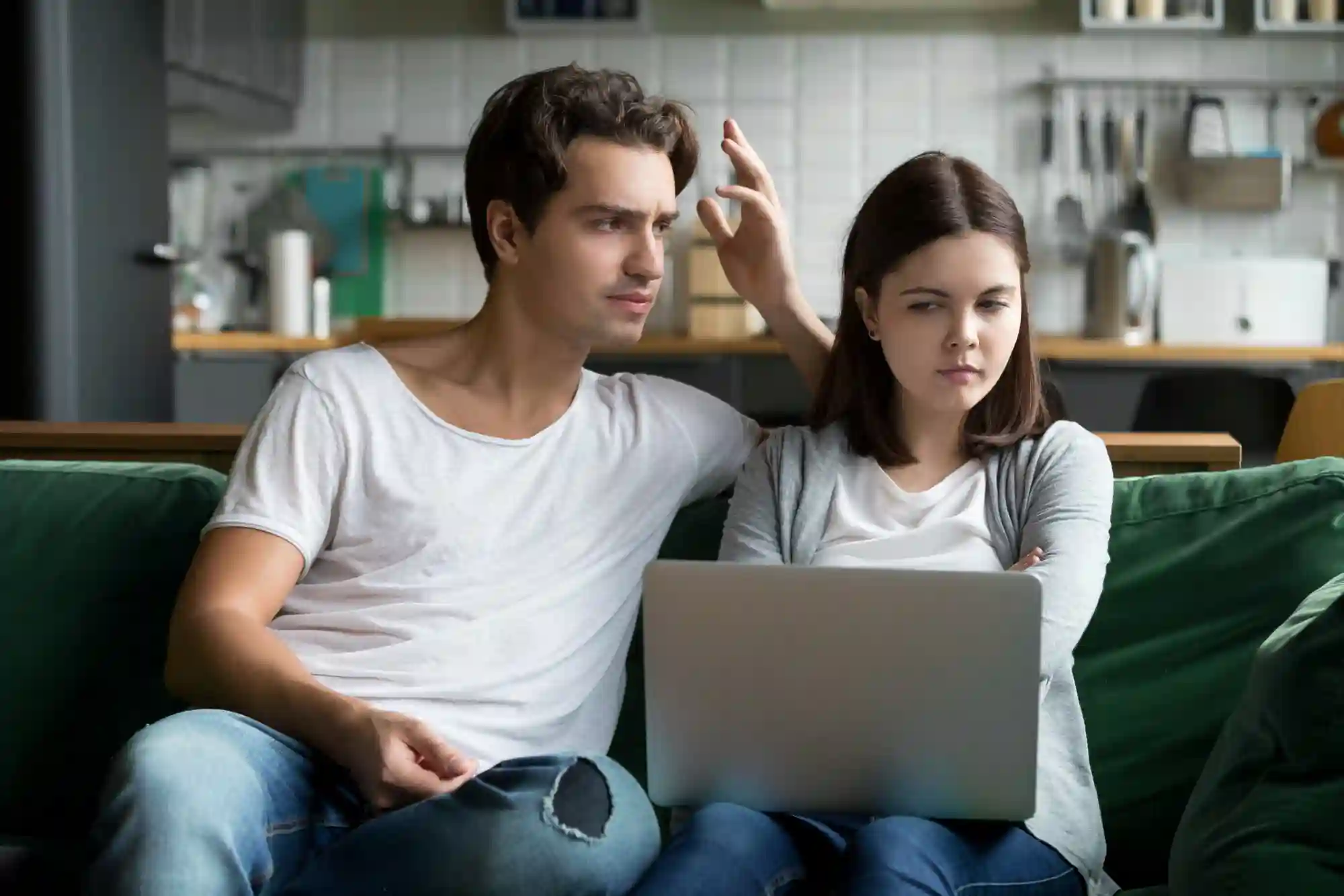 Frustrated young couple arguing on a couch while looking at a laptop, illustrating a common relationship conflict.
