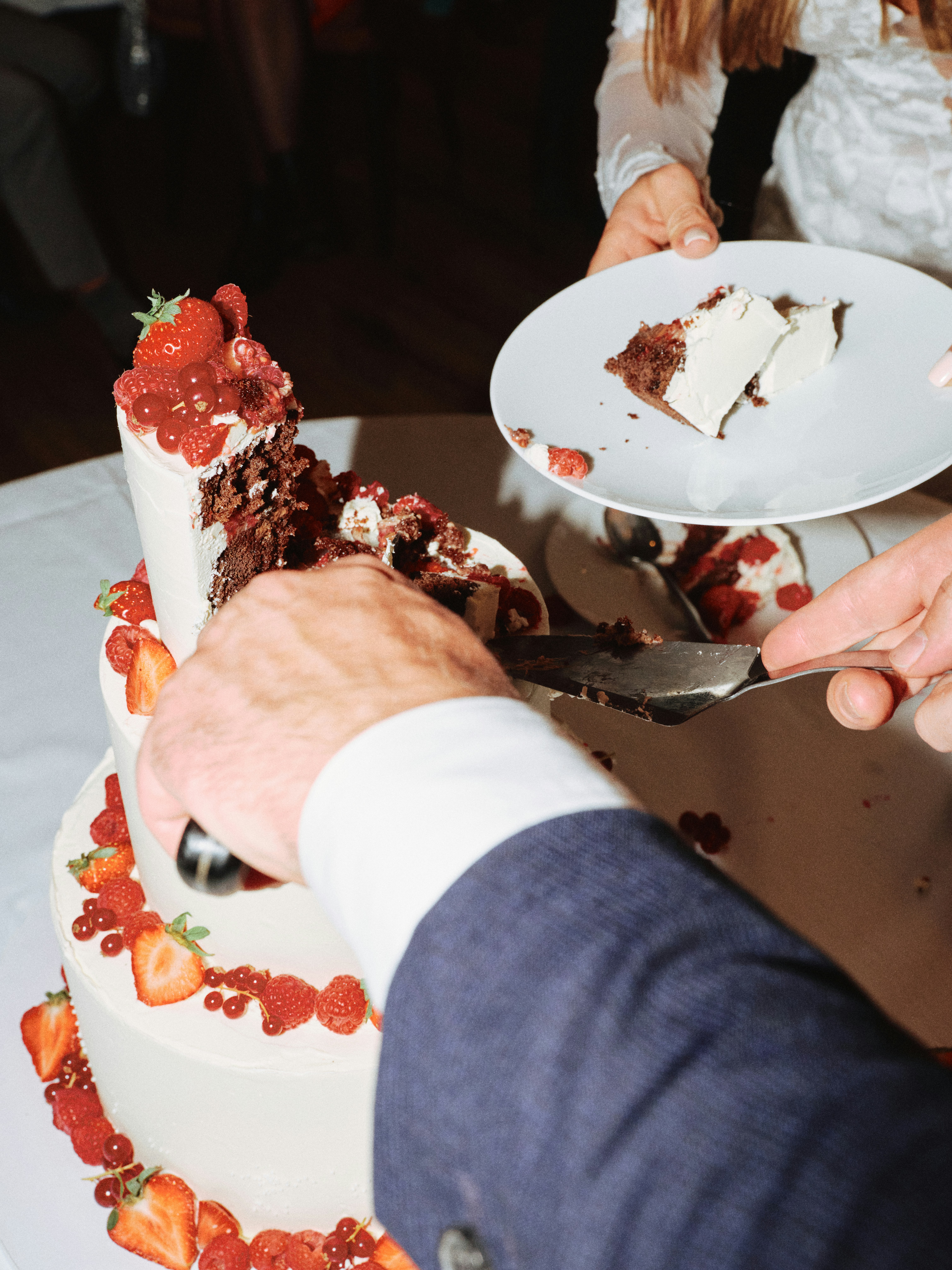 Close-up of a groom's hand serving a slice of chocolate wedding cake onto a white plate, bride's lace sleeve visible in the background