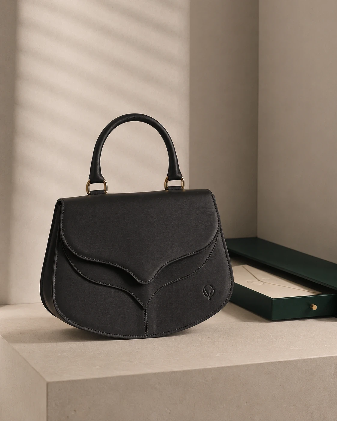 Black leather handbag with a curved flap and gold-tone handle fittings, on a stone pedestal beside a dark green box.