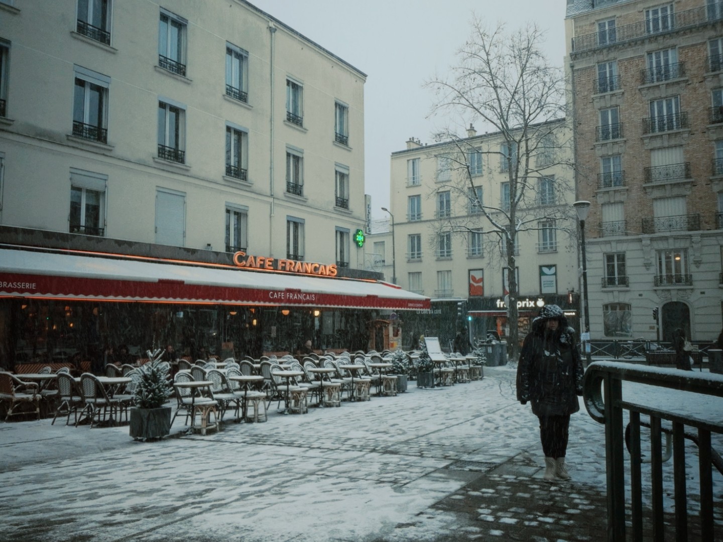 Place enneigée à Issy, devant un Café Français avec terrasse recouverte de neige, personne marchant, bâtiments historiques.