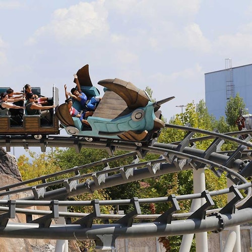 Ihmiset ratsastavat yksityiskohtaisella, lohikäärmeteemaallisella vuoristoradalla aurinkoisena päivänä, taustalla puita ja rakennus.