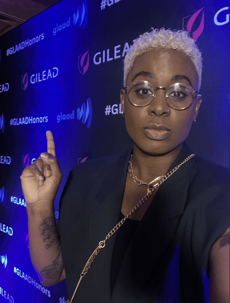 Ashleigh Warren with short blonde hair poses confidently, pointing with one hand at the GLAAD Honors Party, against a blue backdrop with logos.