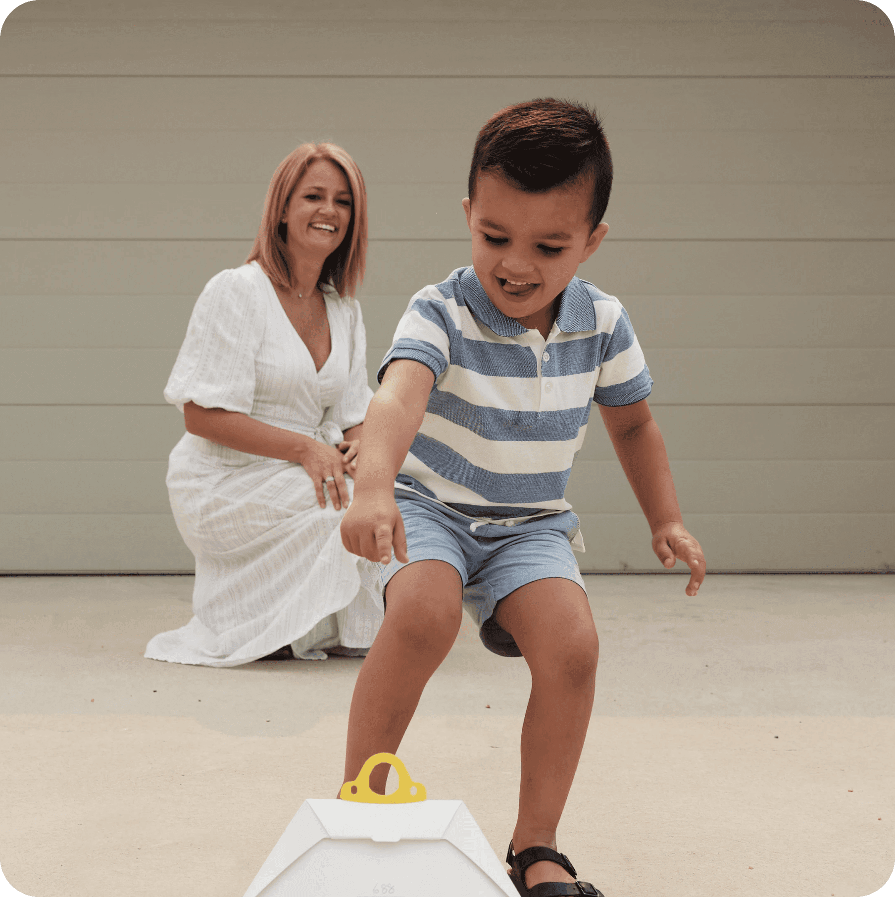 A woman crouches on a driveway in front of a garage door, smiling at a young girl in a peach dress holding a delivery package.