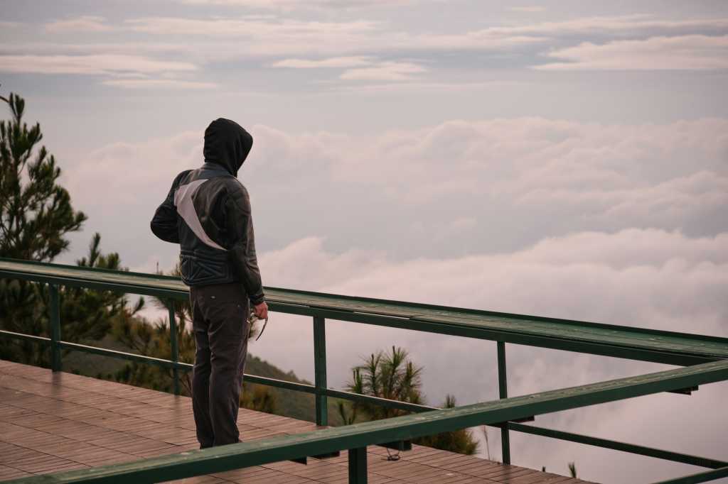 Person in hoodie standing on a platform looking out over clouds