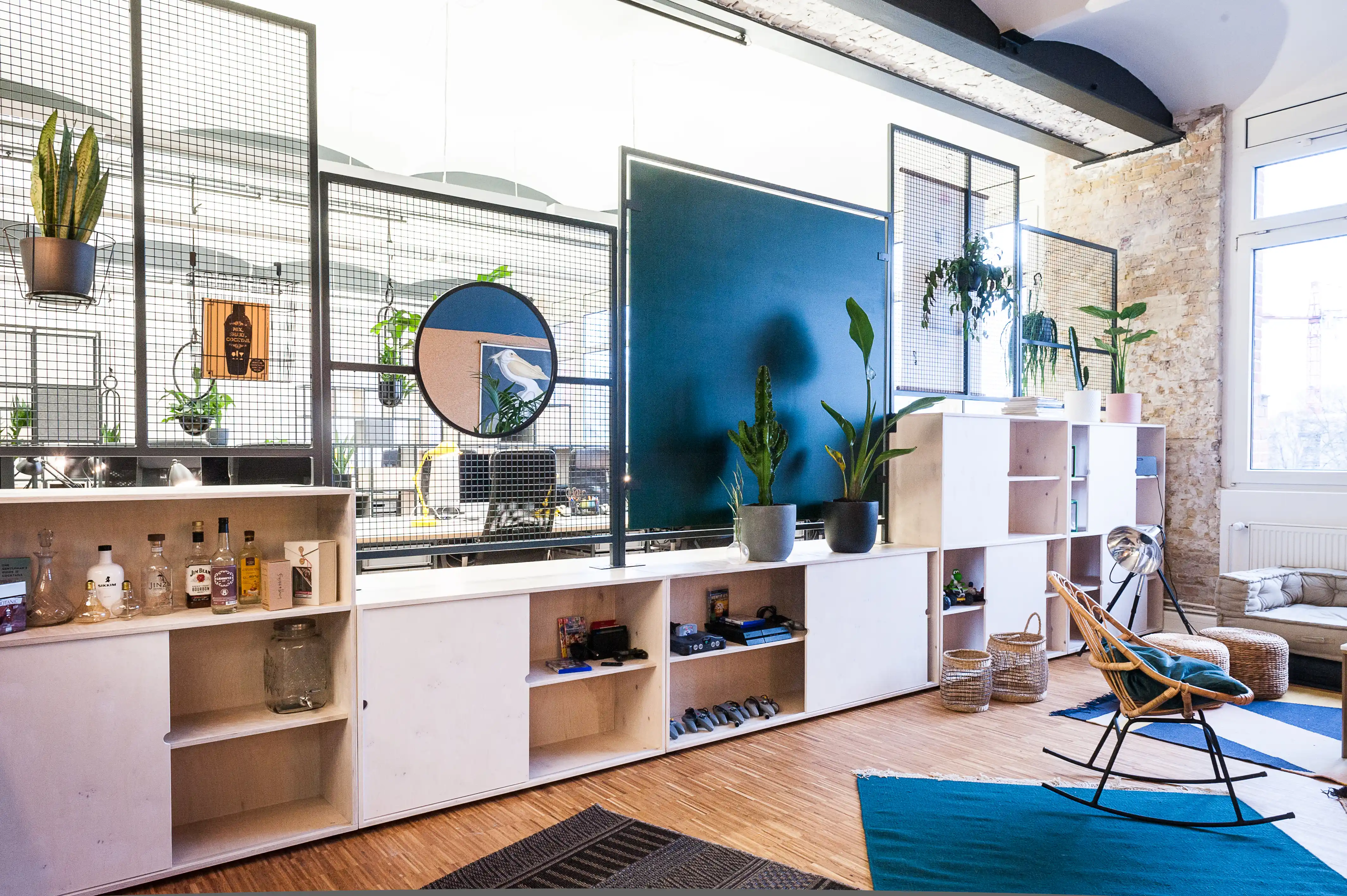 Kitchen bar at INTERMATE x TRUEMATES with birch plywood cabinets holding spirits bottles, black mesh screens with hanging plants, a round mirror, a petrol-blue panel and a rocking chair with teal cushion.