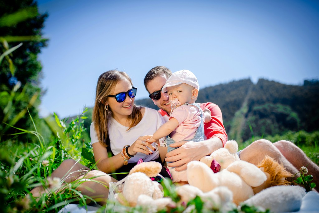 A happy family enjoys a sunny day outdoors, sitting on the grass with their baby and surrounded by plush toys. The image captures the essence of family bonding, outdoor recreation, and the joy of spending quality time together in nature.