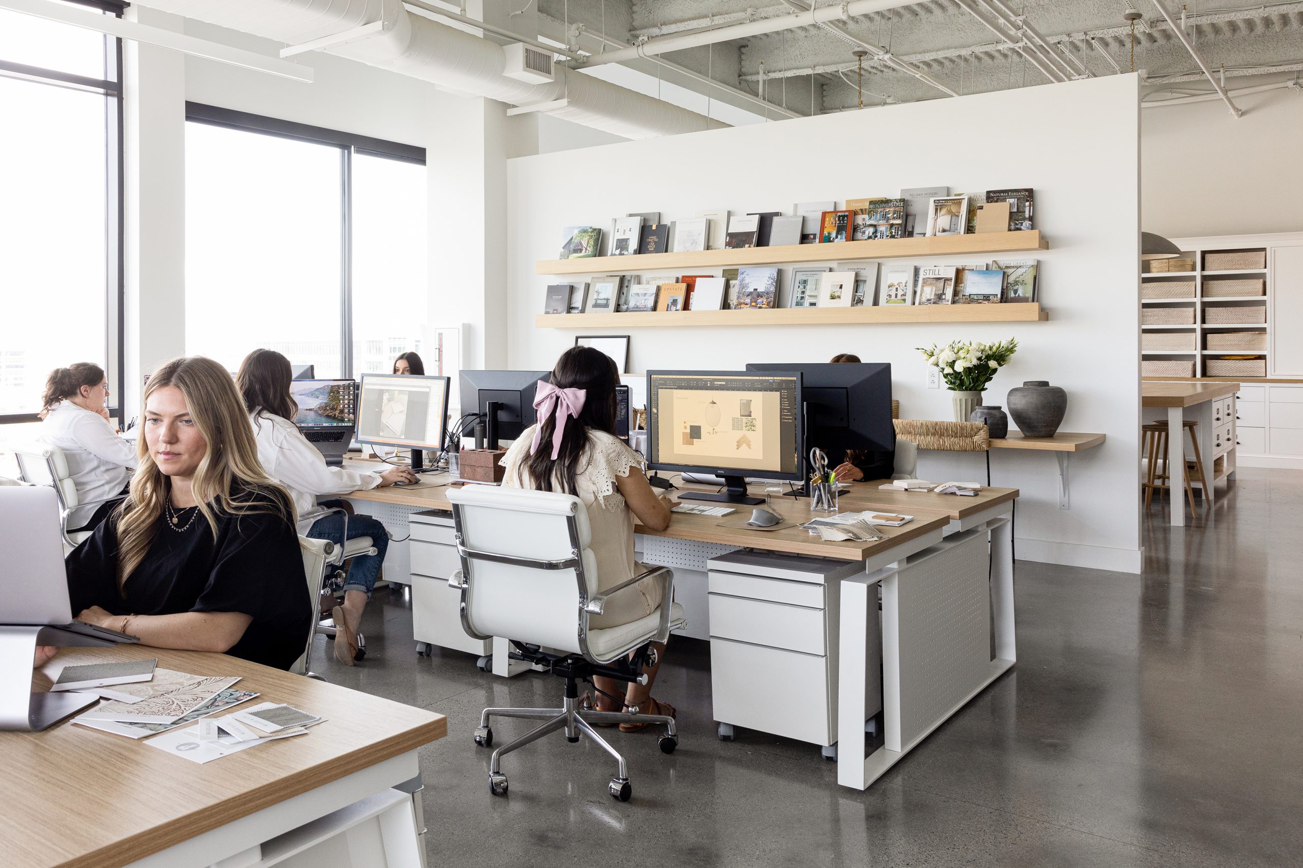 Interior of Studio McGee's office showing team members working at modern workstations with light wood desks and white Eames-style office chairs. The bright, minimalist space features floating shelves displaying design books and magazines, exposed ceiling systems, and polished concrete floors.