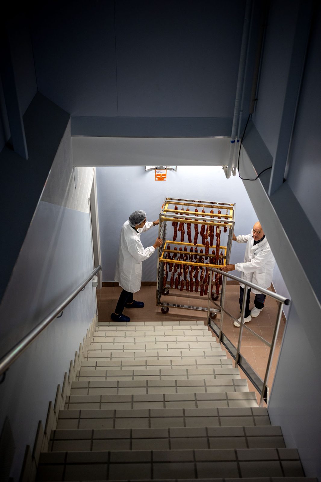 Scala che scende alla cella di Borgo Titol: in fondo due persone in camice spostano un carrello di salami