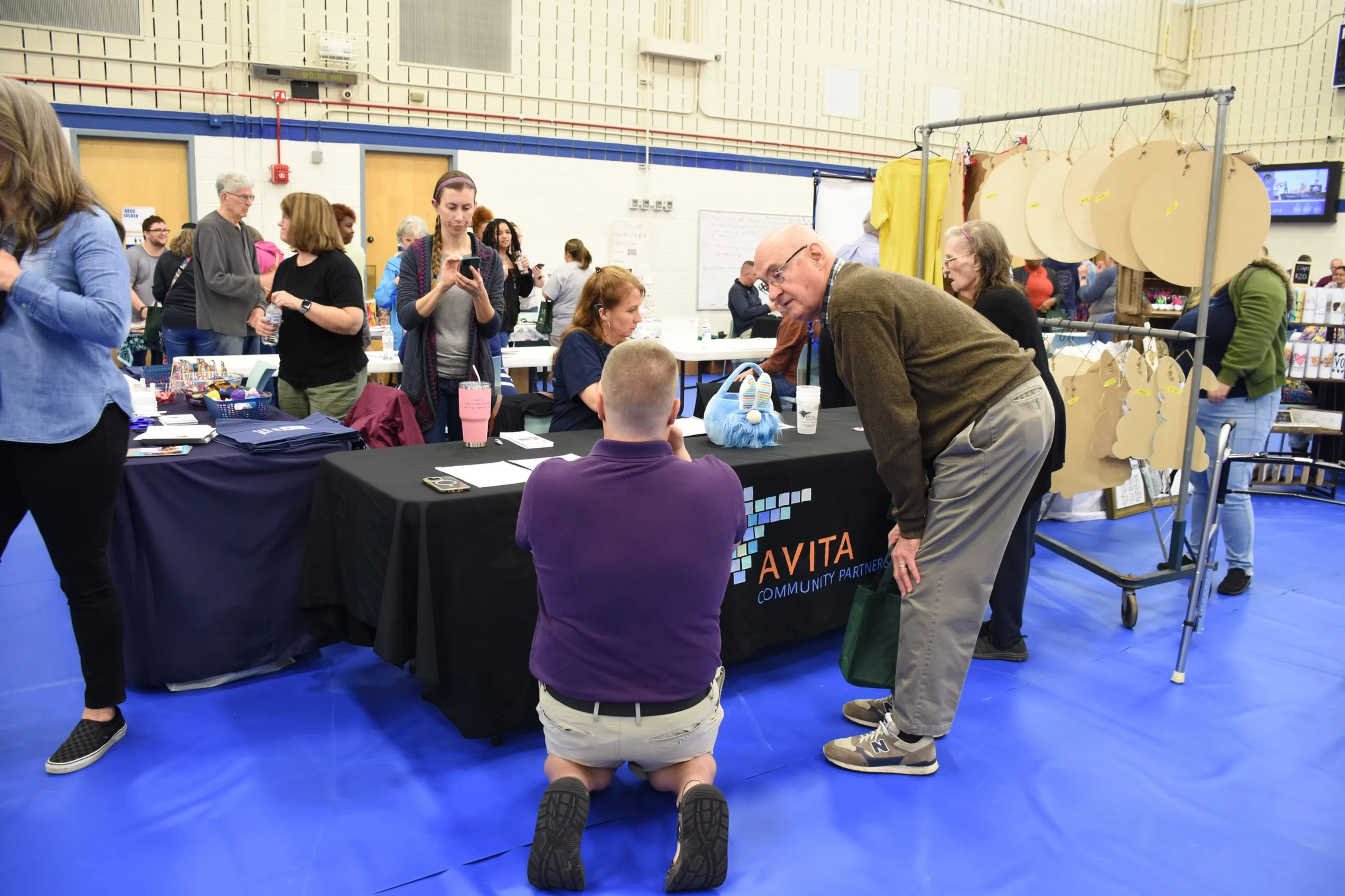 A man kneeling at a table at a crowded event, talking to an older man. Other people are standing around, looking at displays.